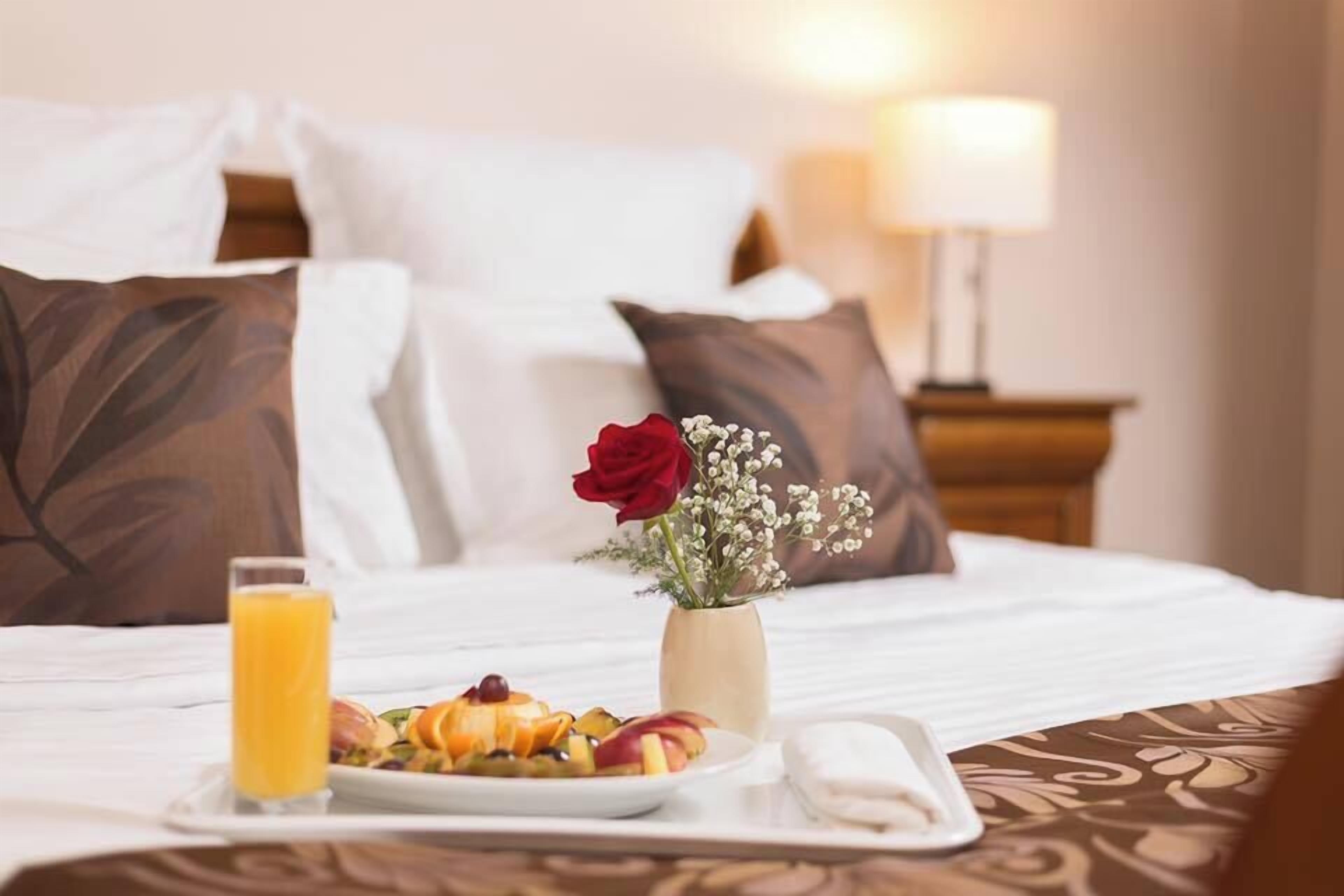 Ein ordentlich bezogenes Bett mit weißen Leinen, ein Tablett mit Frühstück, darunter Obst und ein Glas Orangensaft, eine Vase mit einer roten Rose und Babys Atem, sowie eine Nachttischlampe.