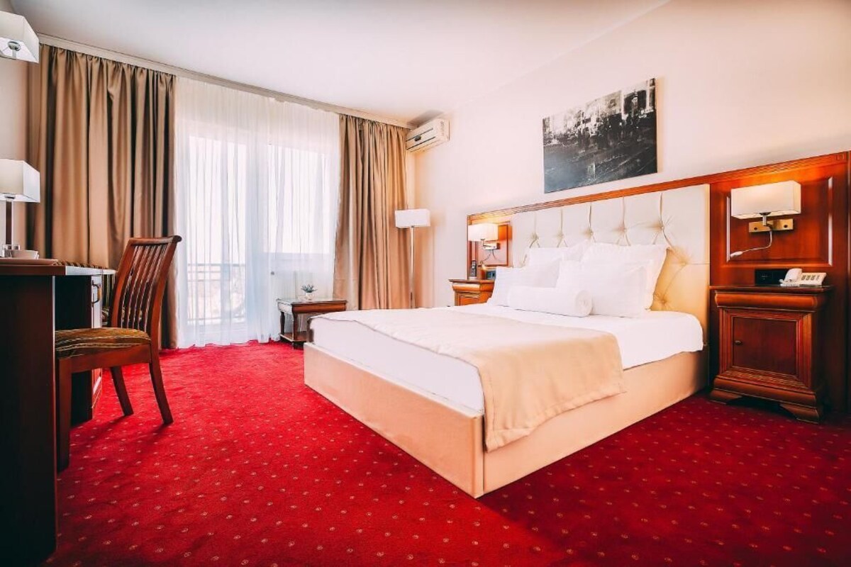 Habitación de hotel con una cama grande, un escritorio y una silla. Hay una ventana con cortinas, un aire acondicionado de pared y un cuadro sobre la cama.