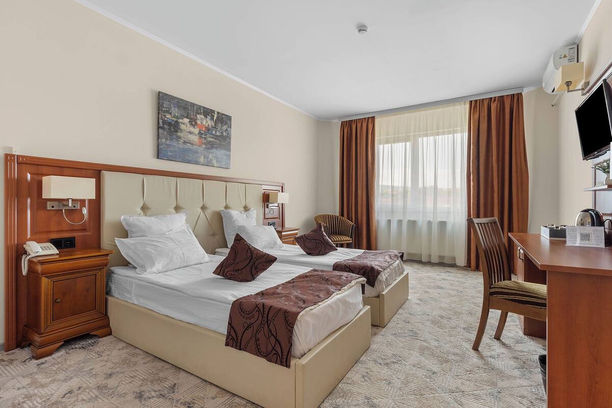 A hotel room with two beds, a desk, a chair, a painting, and a television.