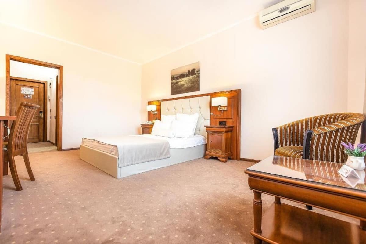 A hotel room with a bed, a wooden nightstand, a chair, a table, and an air conditioning unit.