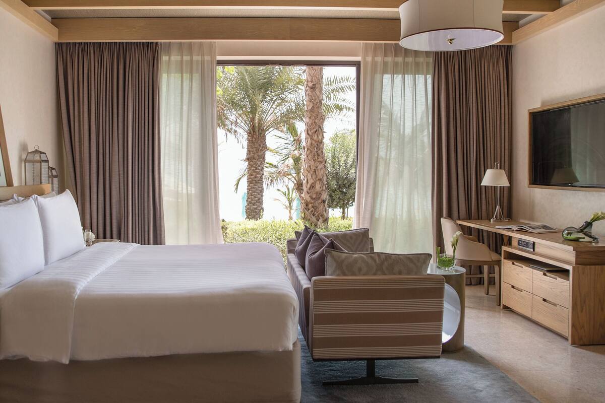 Habitación de hotel con una cama grande, un escritorio con televisor y vistas a palmeras.