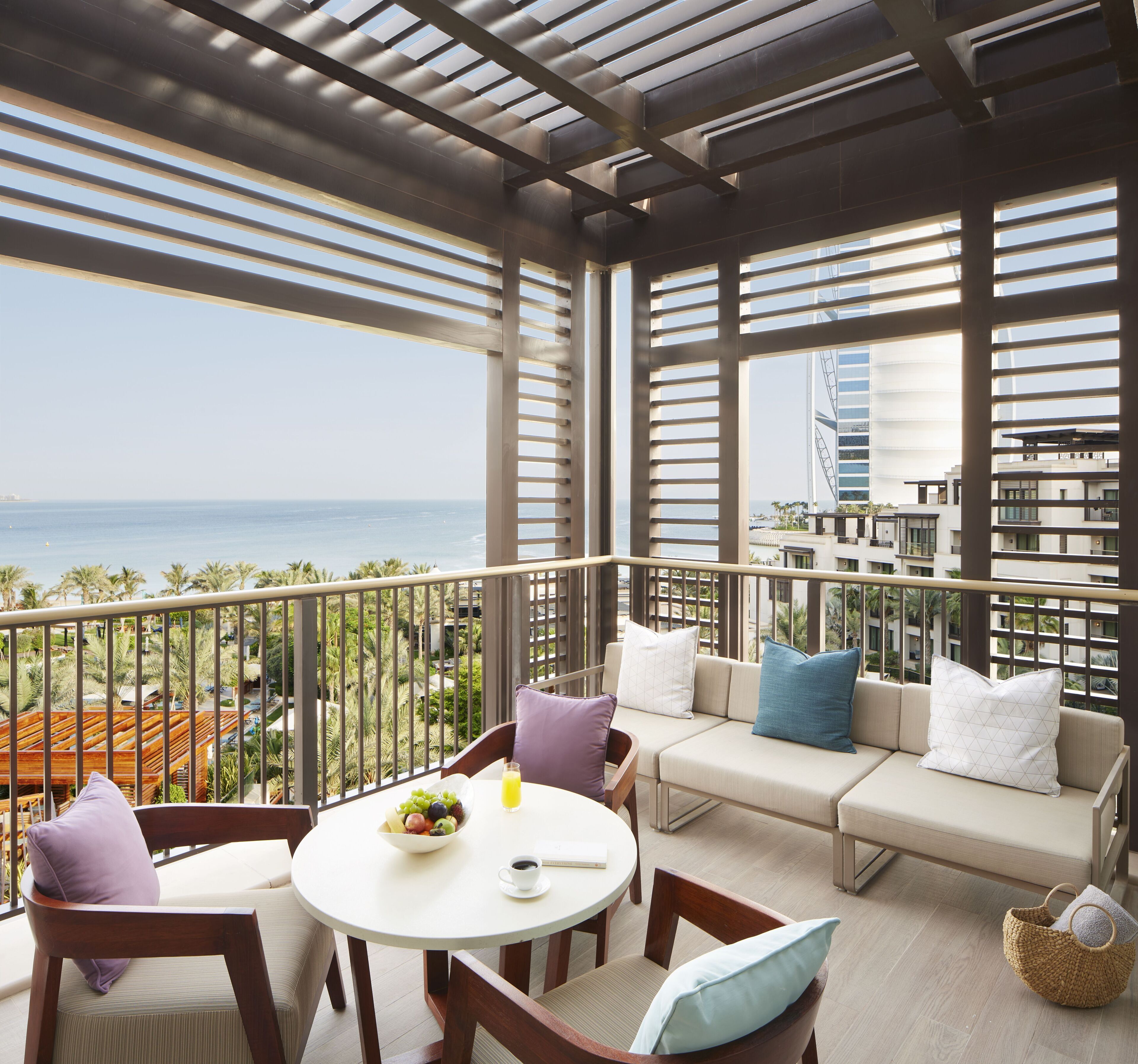 A balcony with a view of the ocean and a cityscape, furnished with a sofa, chairs, and a table with fruit and a drink.