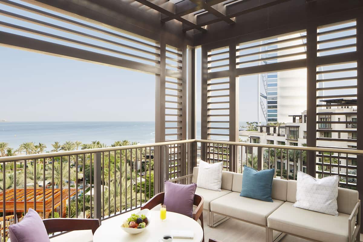 A balcony with a view of the ocean and a cityscape, furnished with a sofa, chairs, and a table with fruit and a drink.