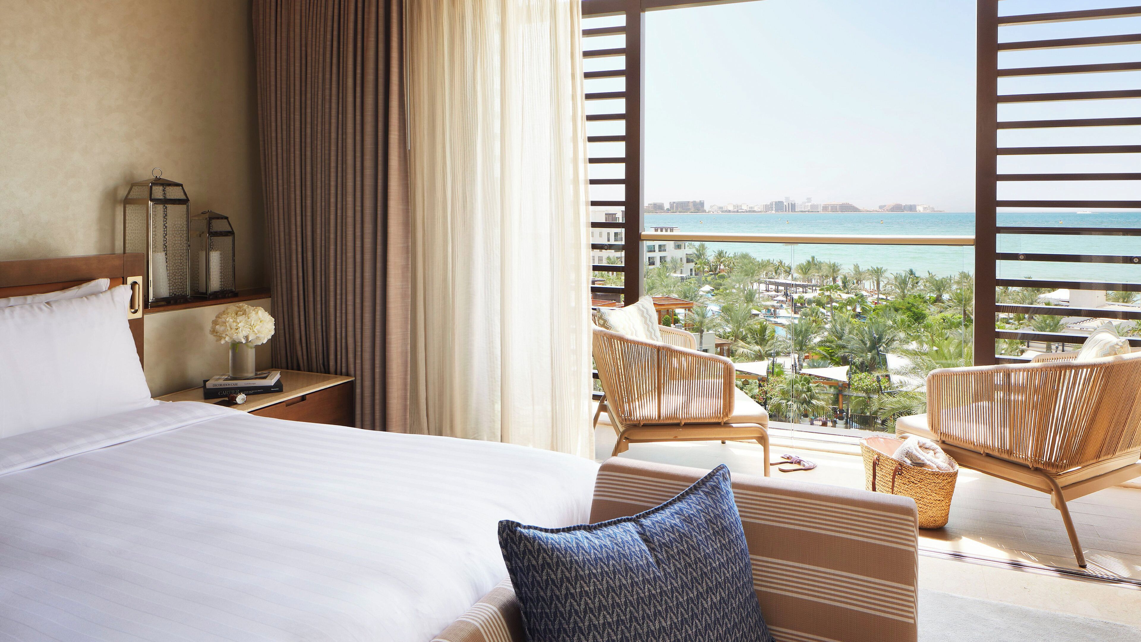 Habitación de hotel con una cama grande, una mesita de noche con una lámpara, un jarrón con flores y vistas a la playa y al paisaje urbano.