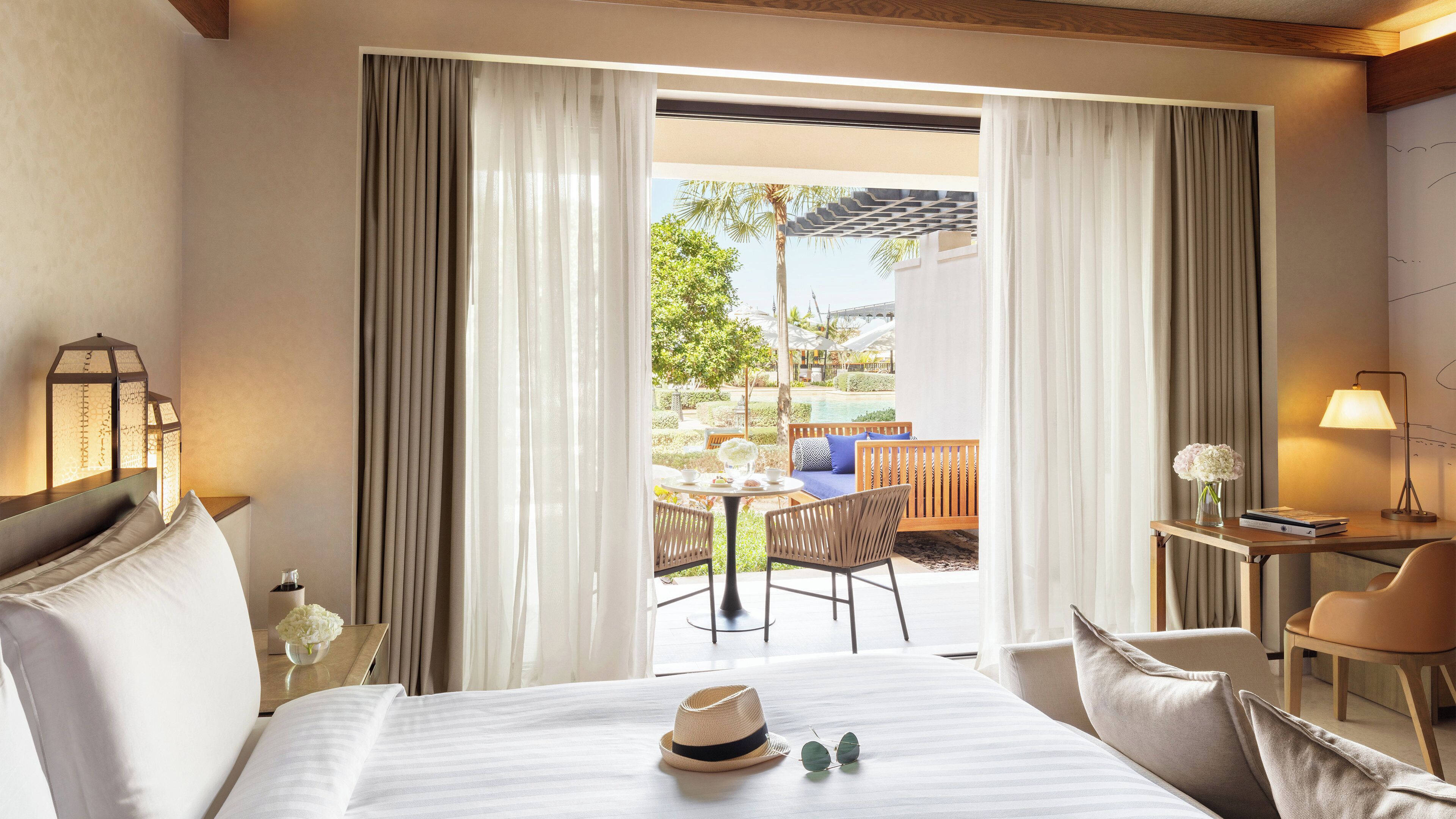 A hotel room with a bed, a desk with a lamp, a chair, and a view of an outdoor area with a pool and greenery.