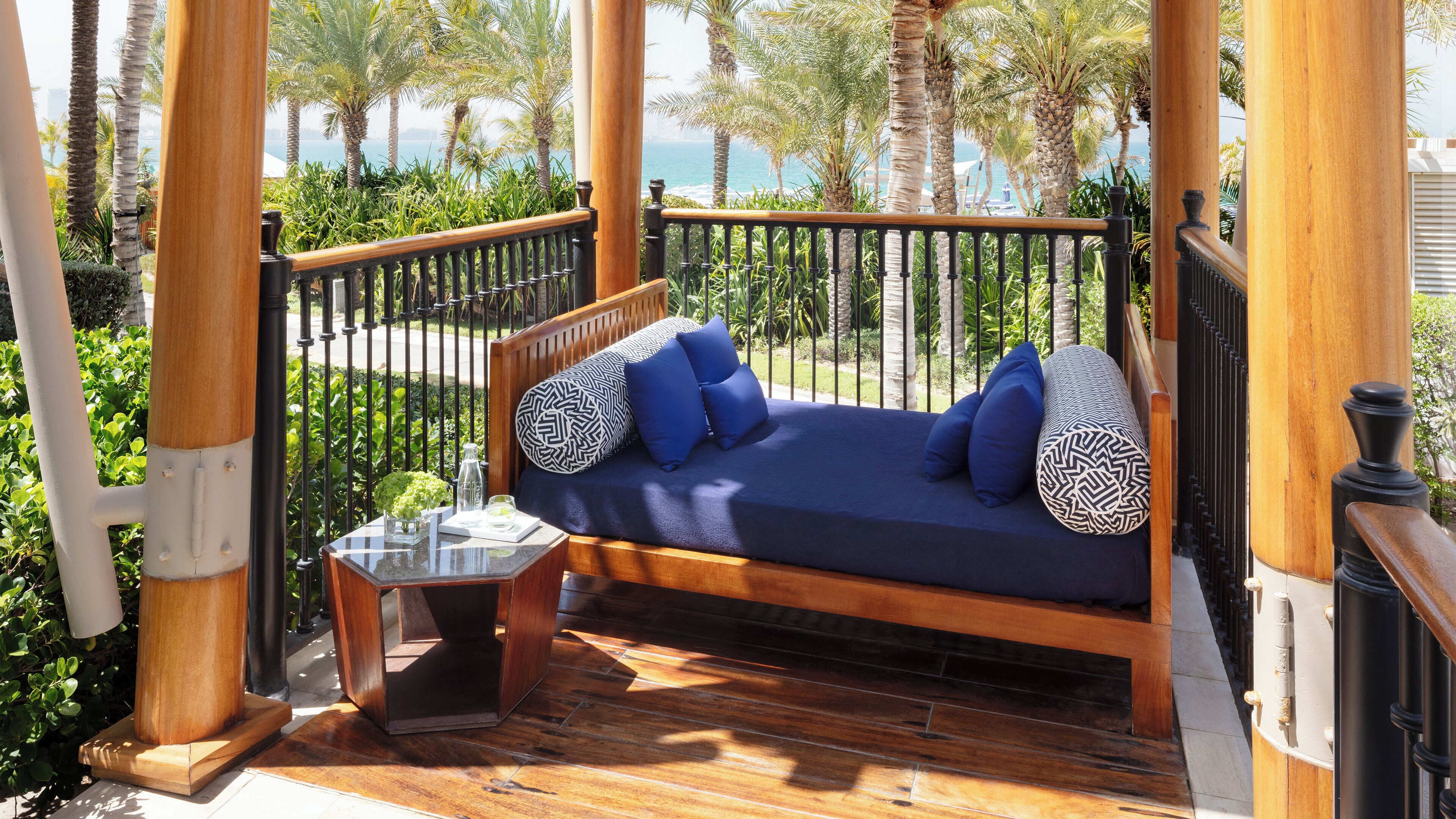 A wooden deck with a sofa and a small table, surrounded by a metal railing and palm trees.