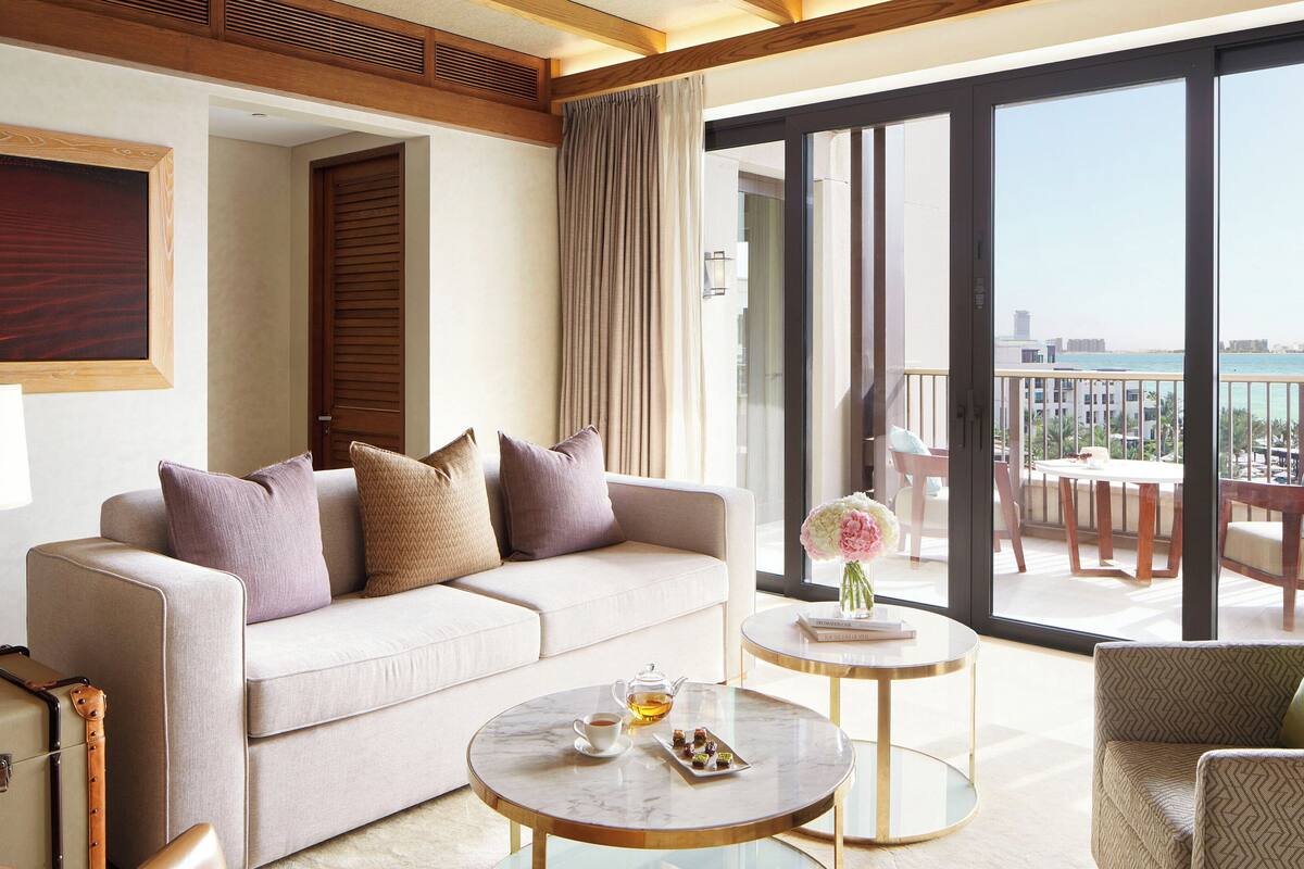 Una habitación de hotel moderna con un sofá, una mesa redonda con tetera y pasteles, vistas al mar y un balcón con mobiliario exterior.