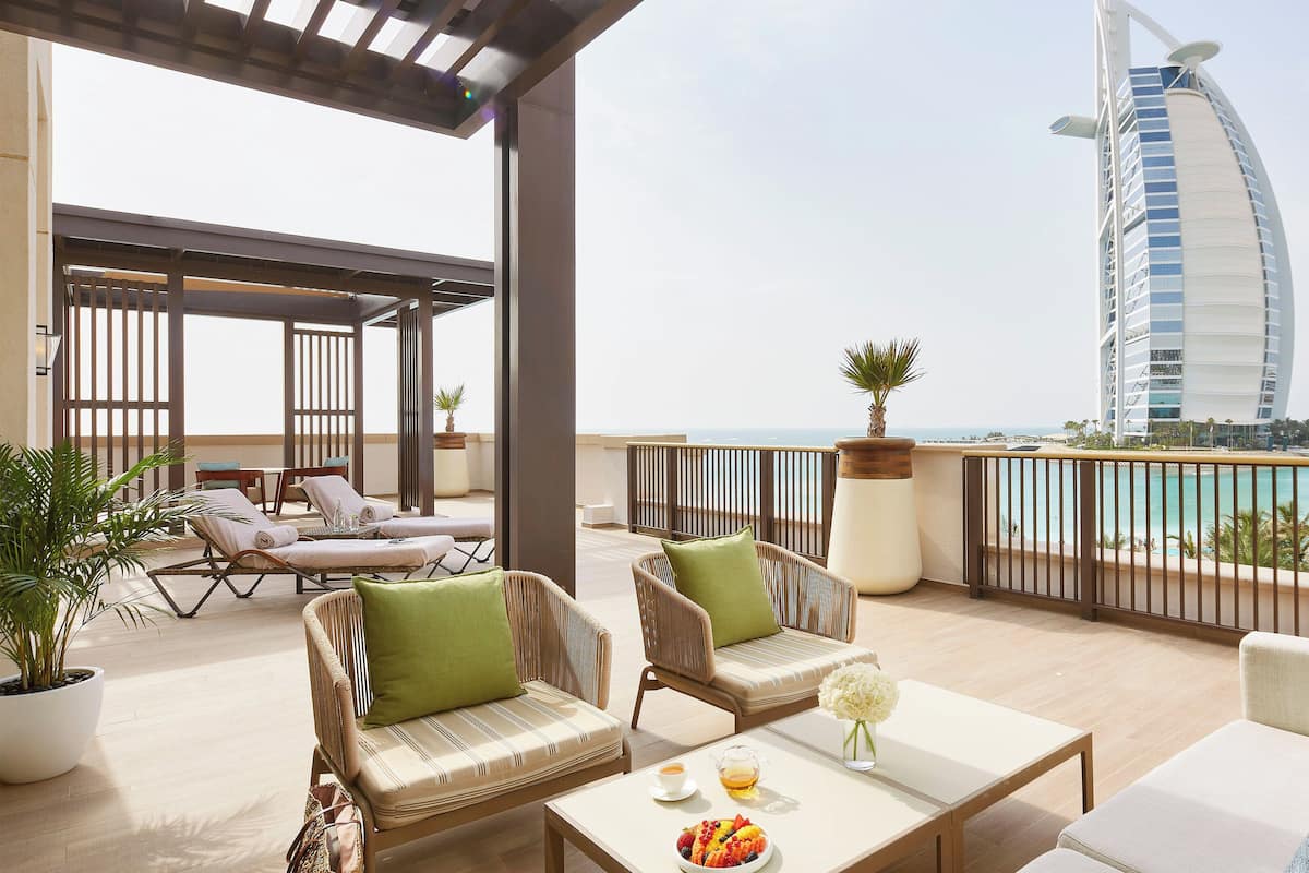 En takterrass med möbler i rotting, ett bord med en fruktskål och drycker, och utsikt över Burj Al Arab-hotellet och havet.