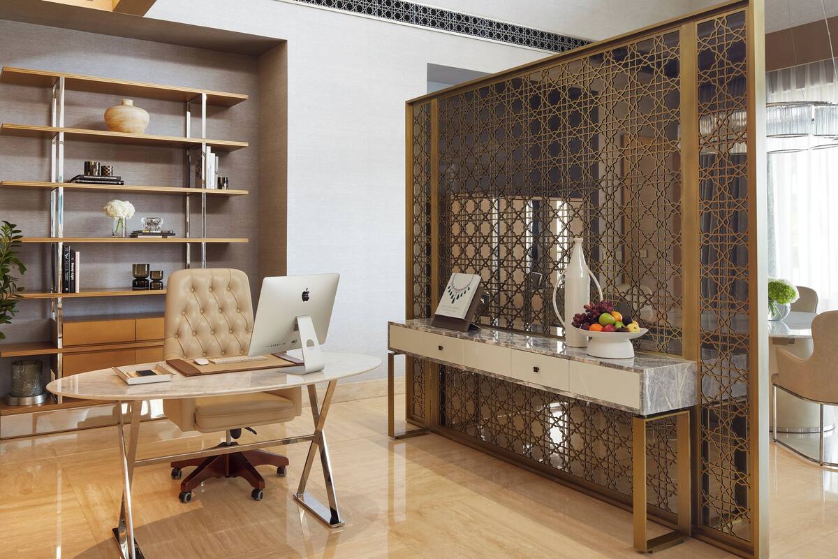A modern office space with a desk, chair, and computer. There are shelves with books and decorative items, and a decorative partition with a geometric pattern.