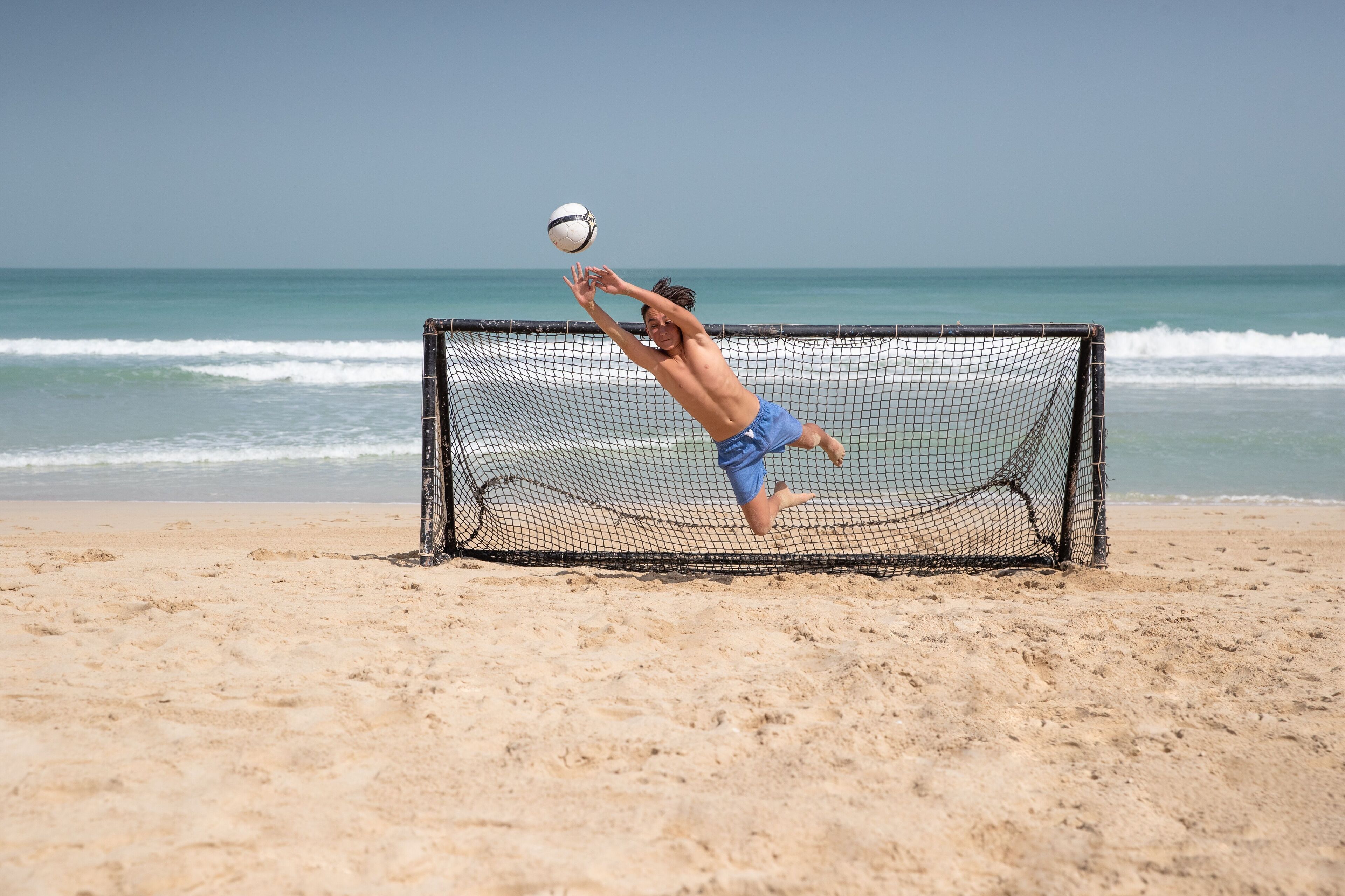 Un joueur de volleyball de plage, vêtu d’un short bleu, saute pour frapper une balle près d’un filet de volleyball.