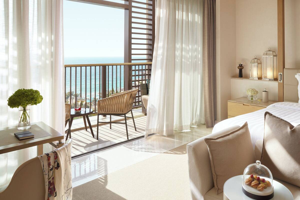 Habitación de hotel con balcón, cama, escritorio con un jarrón de flores y zona de comedor con mesa y sillas.