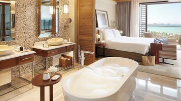 A modern hotel room with a large bathtub, a wooden vanity, a large mirror, and a bed with a view of the sea.