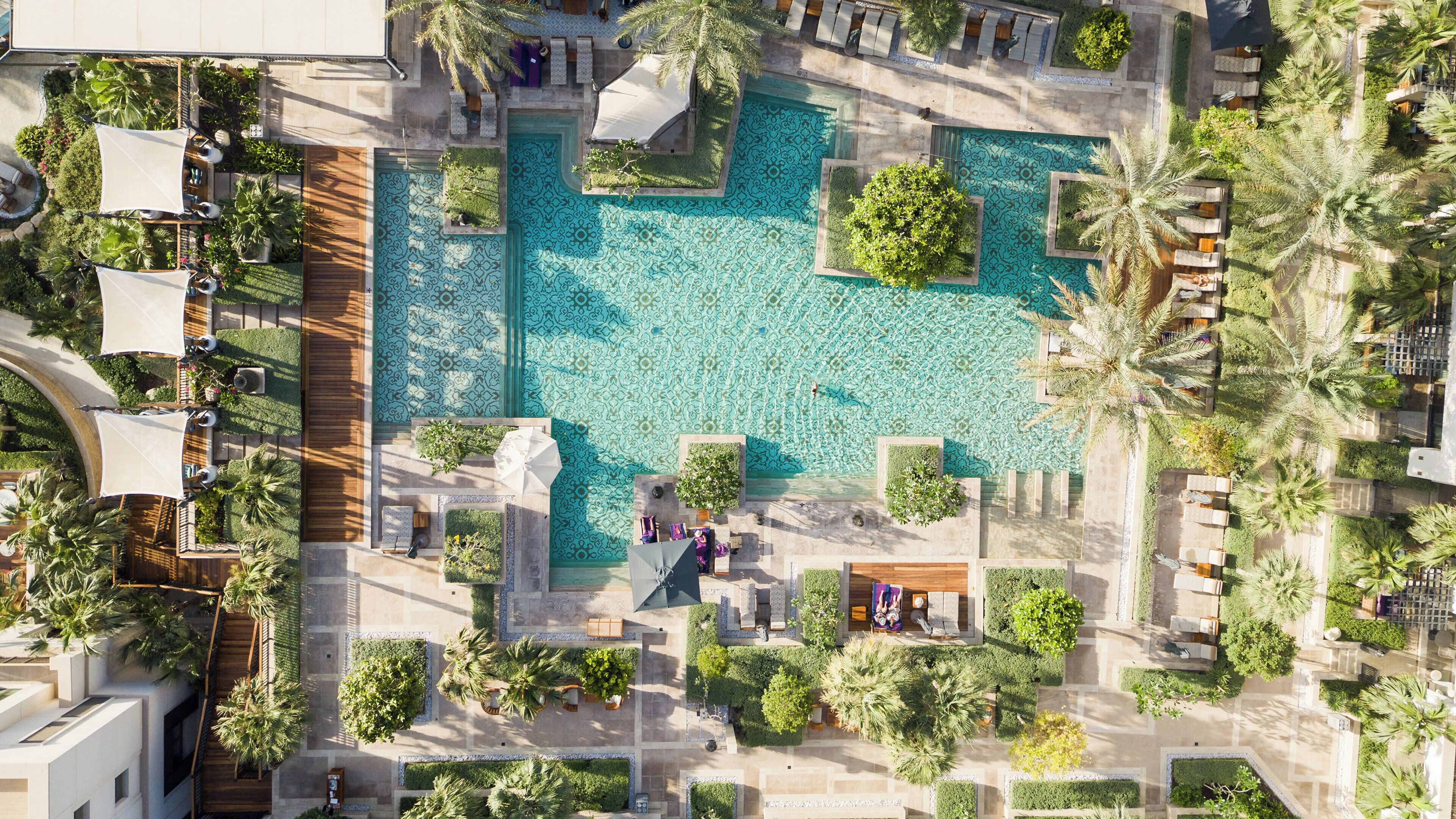Eine Vogelperspektive auf ein Resort mit einem zentralen Schwimmbecken, umgeben von Liegebereichen, Palmen und einem gepflegten Garten.