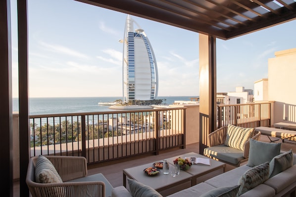 A balcony with a view of a distinctive sail-shaped building, wicker furniture, and a table with fruits and a pitcher.
