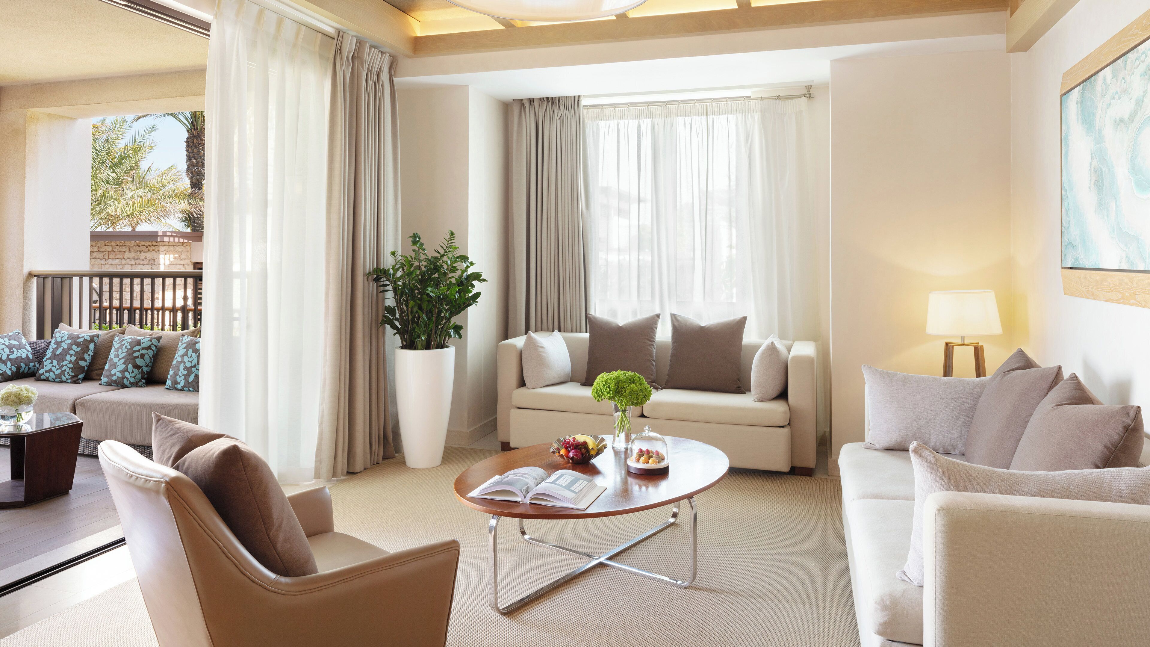 A modern hotel room with a beige sofa, a round coffee table with a book and fruit, a potted plant, and a balcony with a view of palm trees.