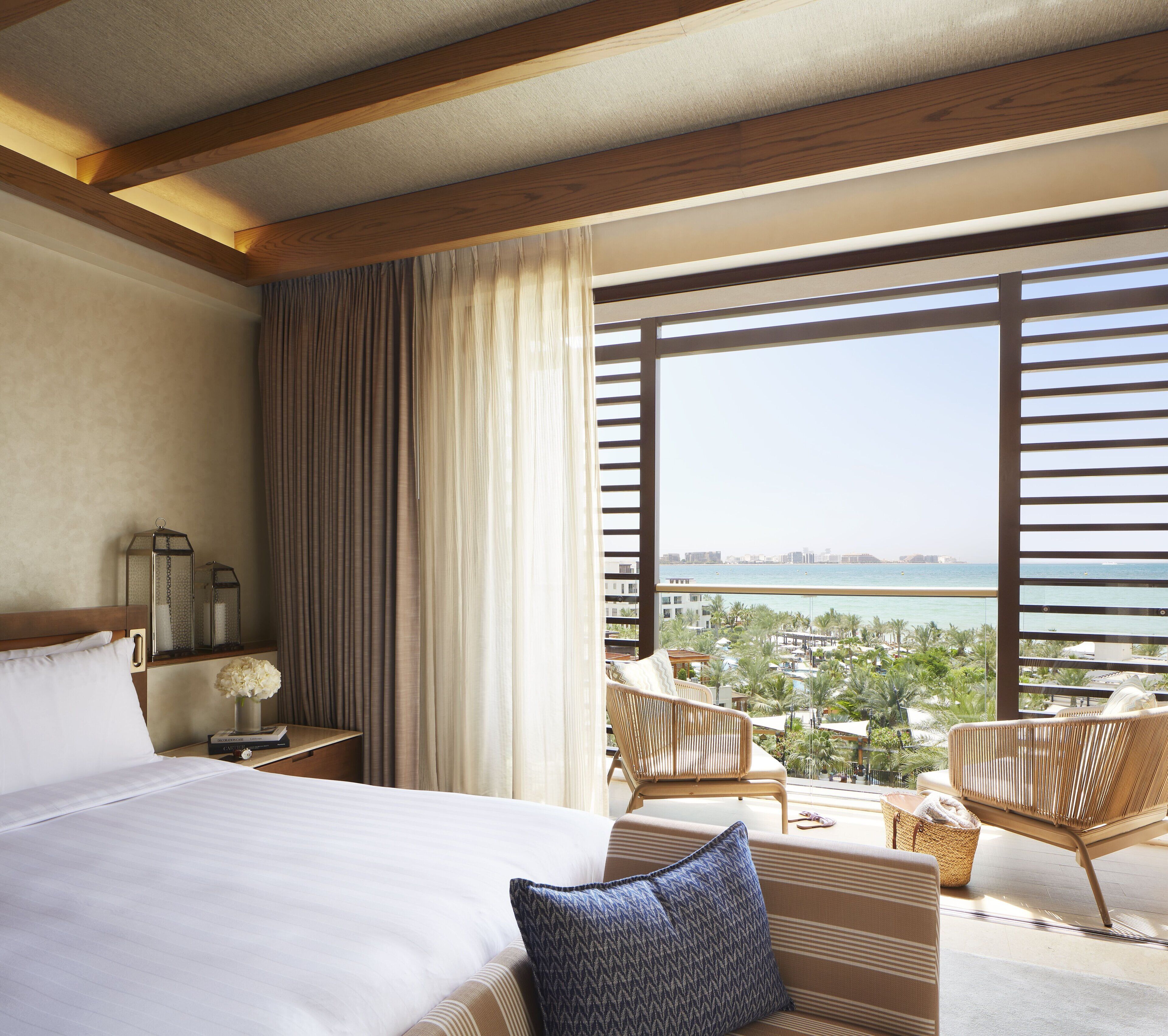 A hotel room with a large bed, a view of a resort and ocean, and a balcony with wicker furniture.