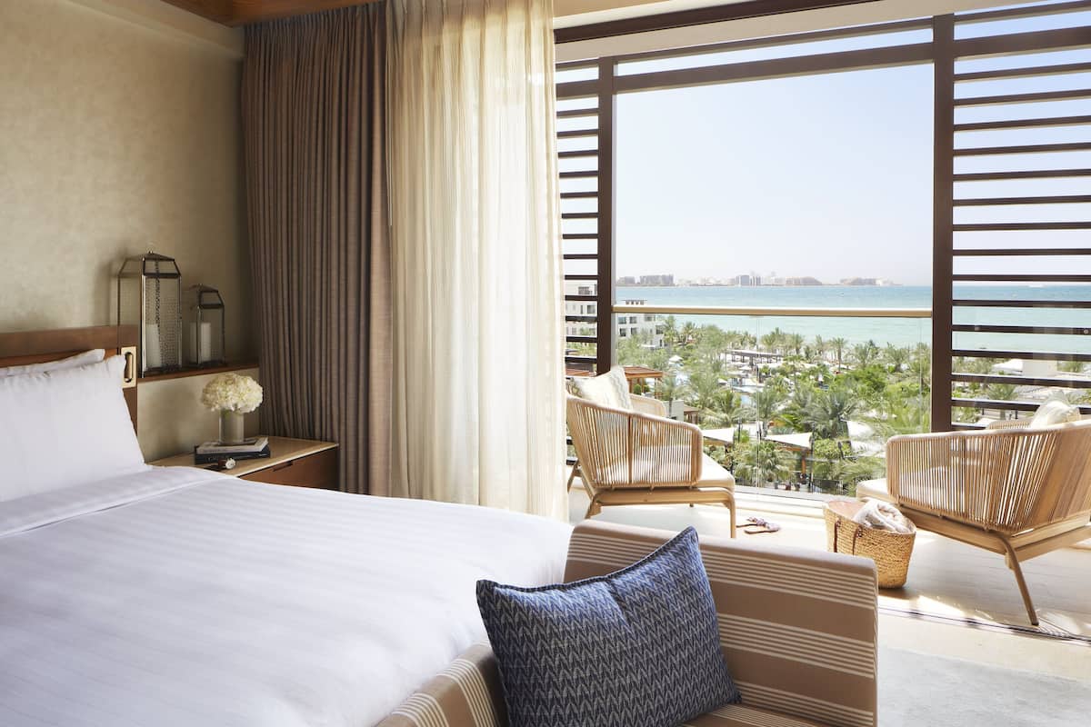 A hotel room with a large bed, a view of a resort and ocean, and a balcony with wicker furniture.