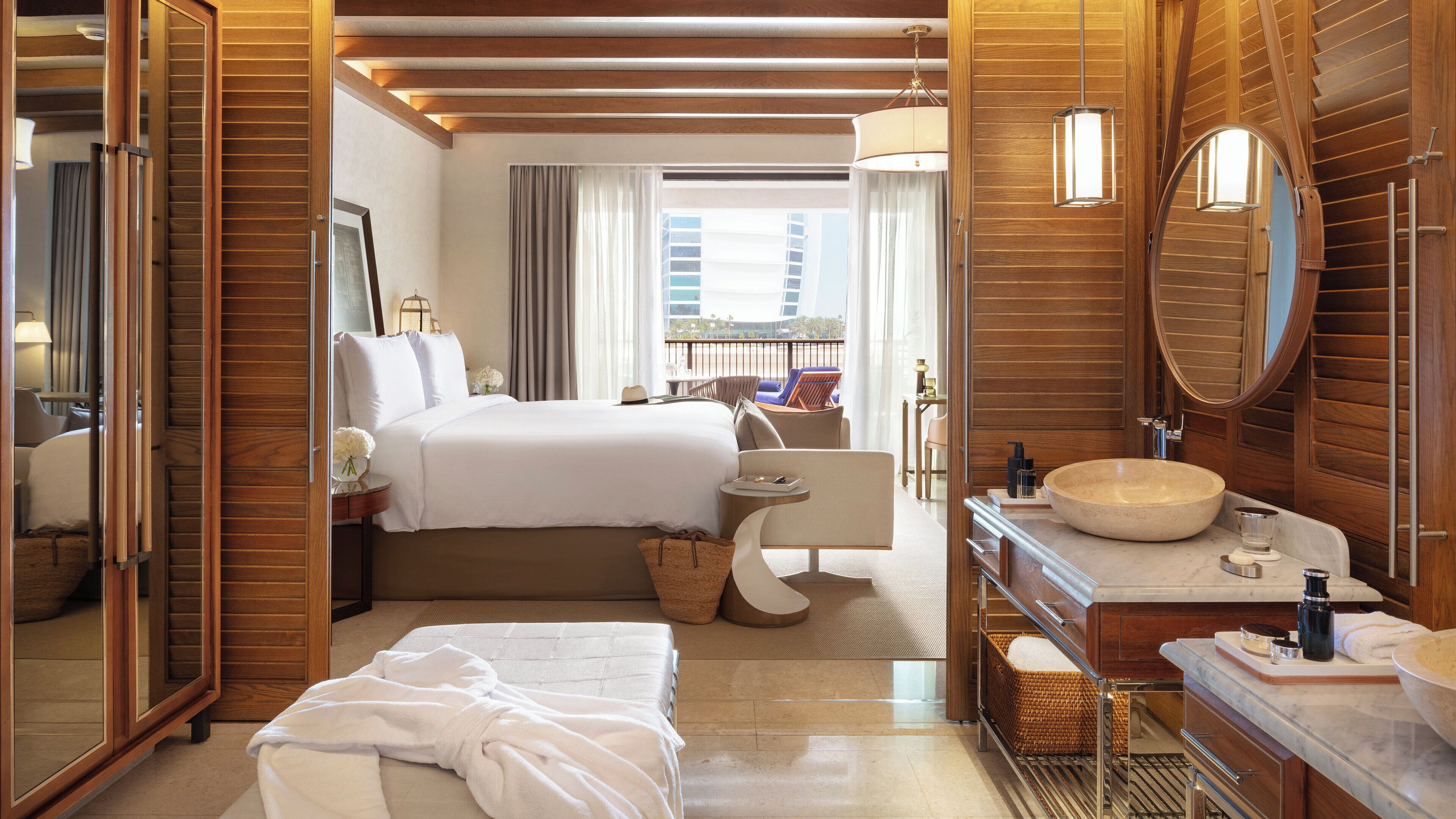 A hotel room with a large bed, a sitting area, a bathroom with a large sink, and a view of the city.