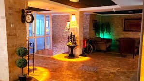 Un hall d’entrée avec un sol carrelé, une horloge murale, une lampe décorative et une porte donnant sur une autre pièce.