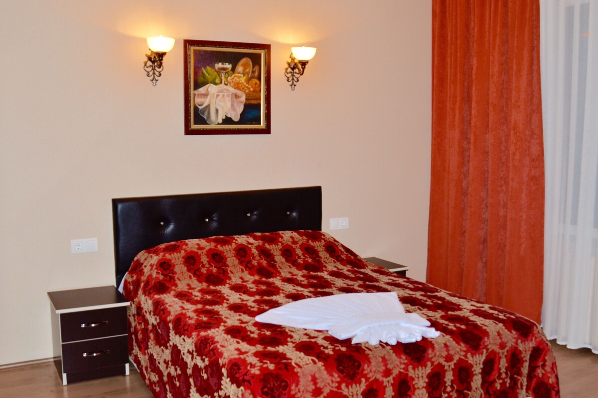 A bedroom with a bed, a nightstand, a wall-mounted painting, and a window with curtains.
