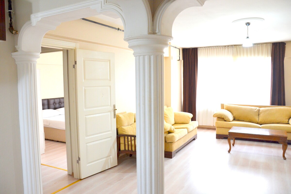 A room with a yellow sofa, a wooden coffee table, and a bed in the background.