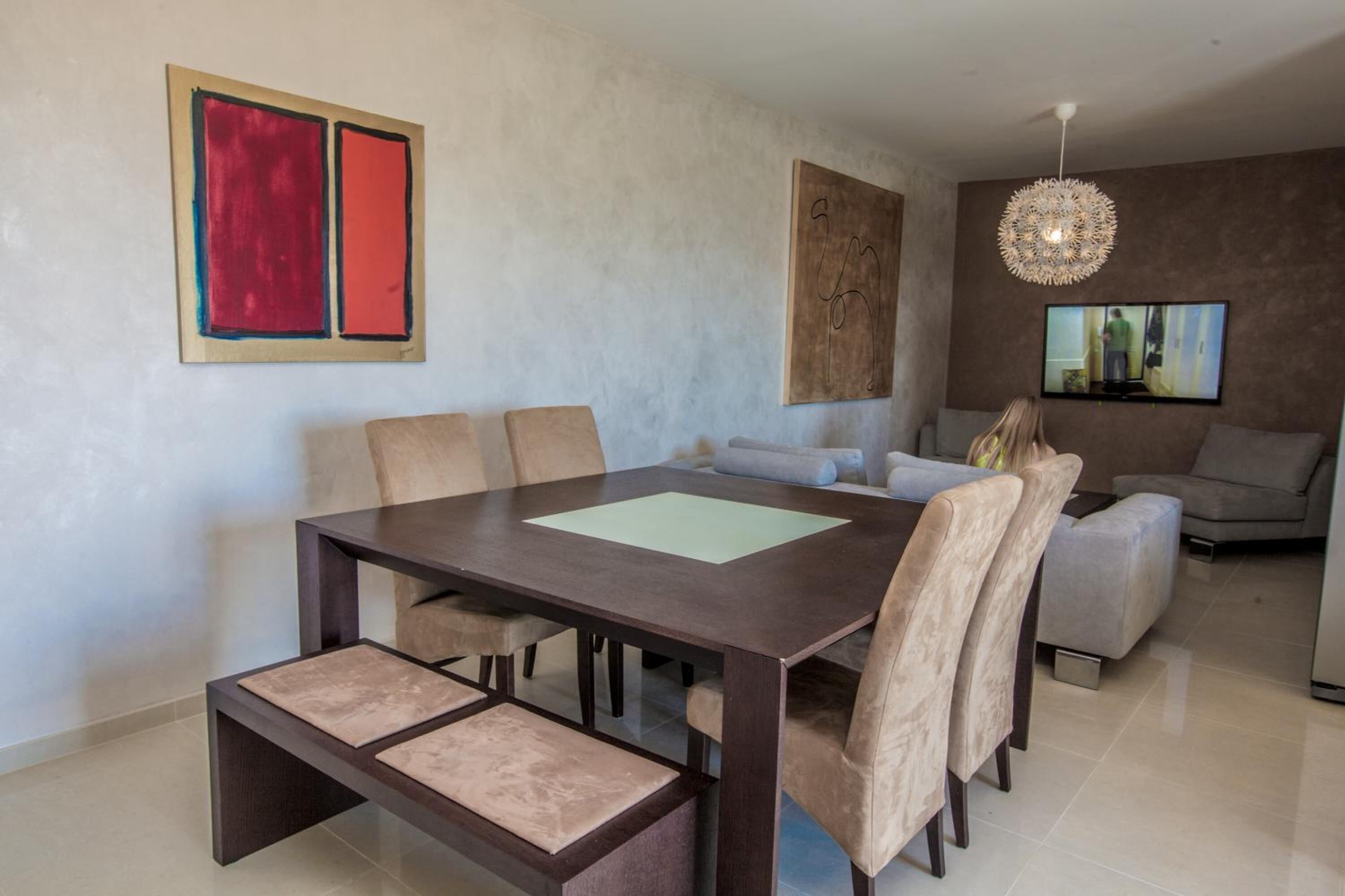 Un espace repas moderne avec une table en bois foncé et des chaises de couleur beige, un canapé et un téléviseur en arrière-plan.