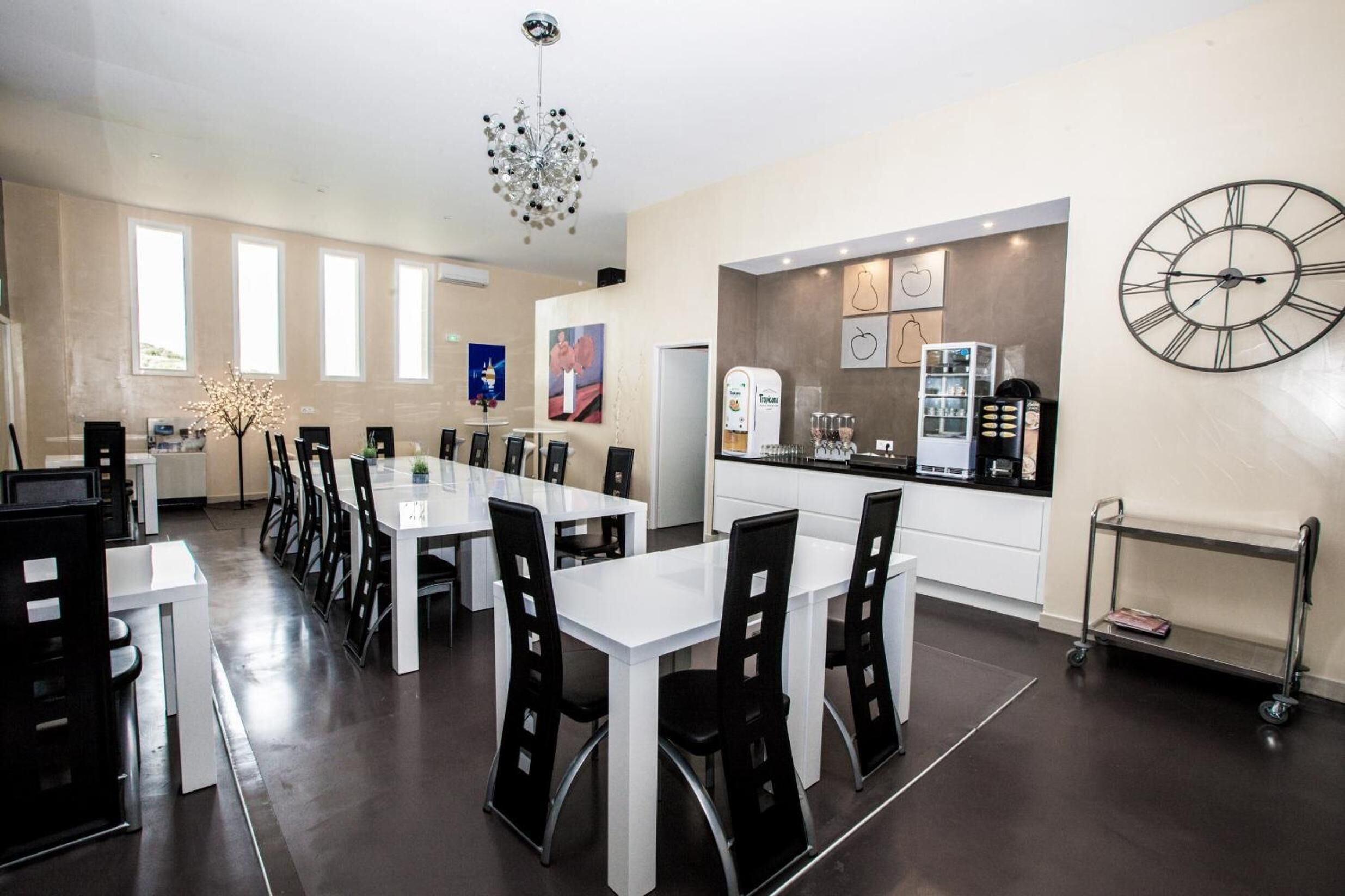 A modern dining area with white tables and black chairs, a kitchen with a clock, and a chandelier.