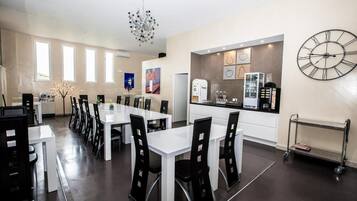 A modern dining area with white tables and black chairs, a kitchen with a clock, and a chandelier.