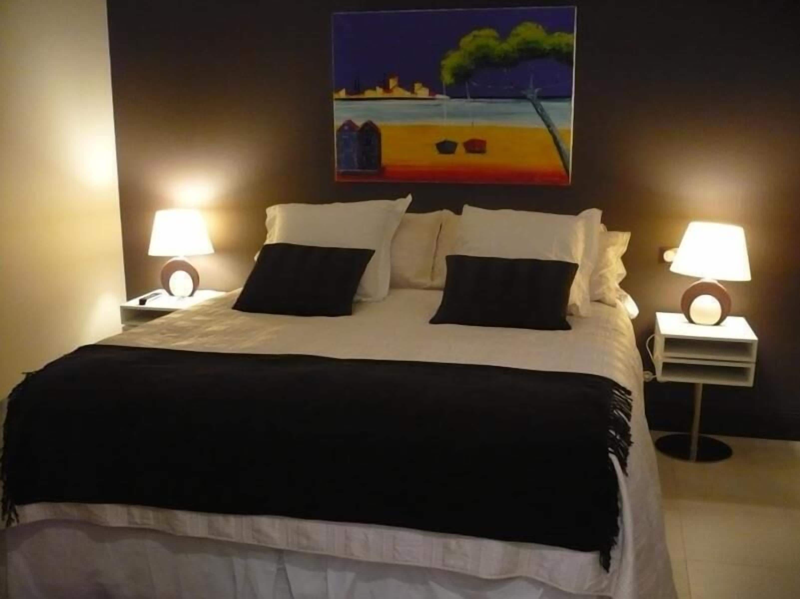 A neatly made bed with white and black bedding, two bedside lamps, and a painting of a beach scene on the wall.
