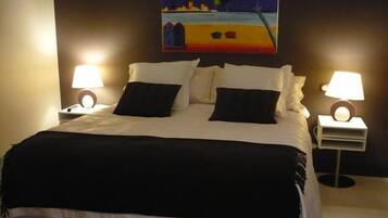 A neatly made bed with white and black bedding, two bedside lamps, and a painting of a beach scene on the wall.