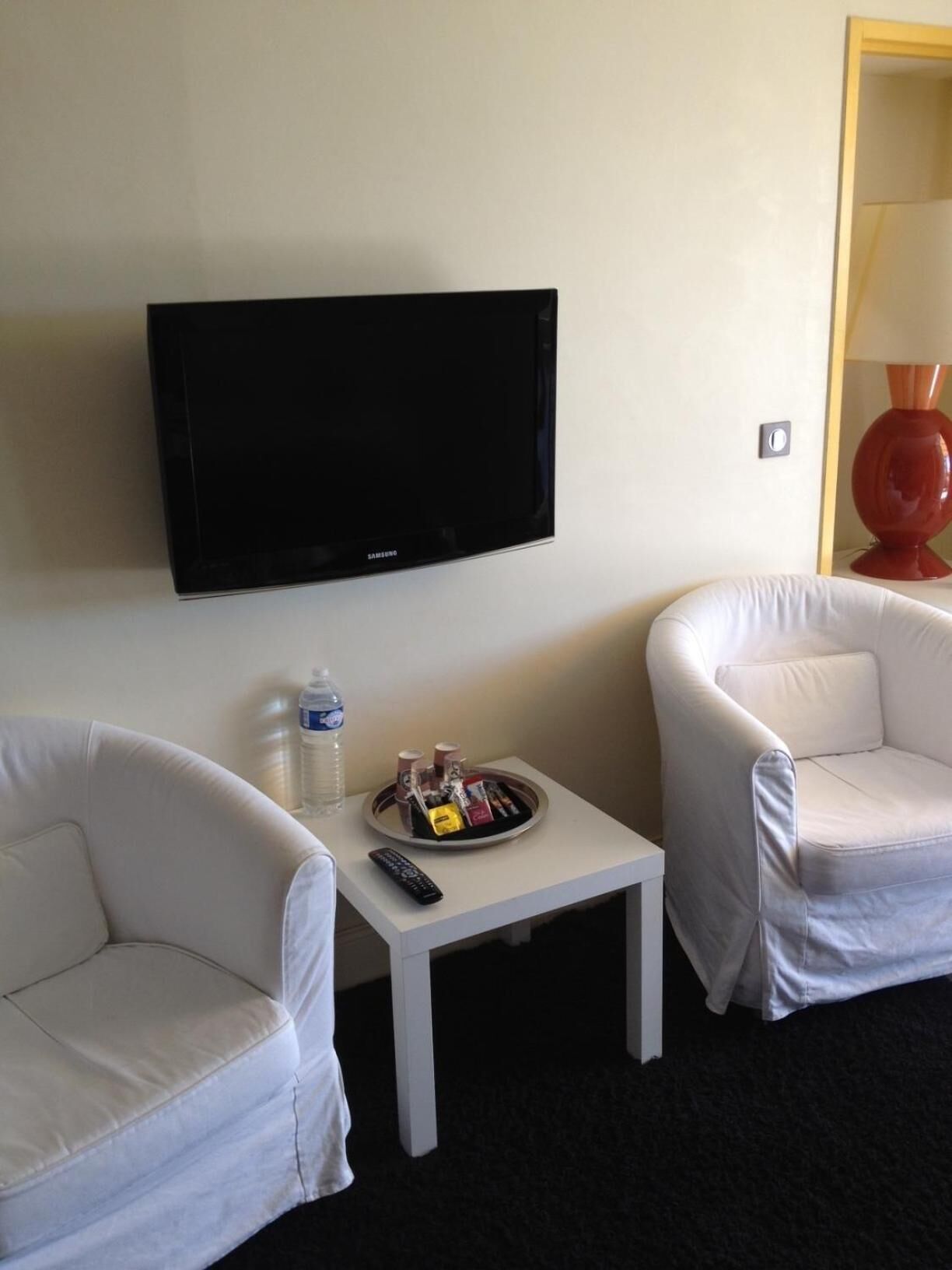 A hotel room with a flat-screen TV mounted on the wall, two white armchairs, a small white table with a tray of snacks and a bottle of water, and a lamp on the right side.