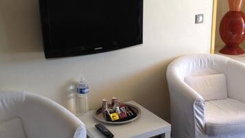 A hotel room with a flat-screen TV mounted on the wall, two white armchairs, a small white table with a tray of snacks and a bottle of water, and a lamp on the right side.