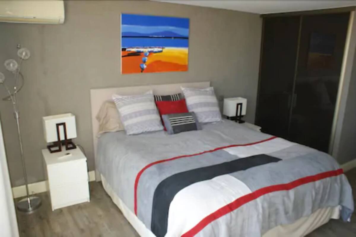 A bedroom with a bed, bedside tables, a nightstand, a wall-mounted lamp, and a painting.