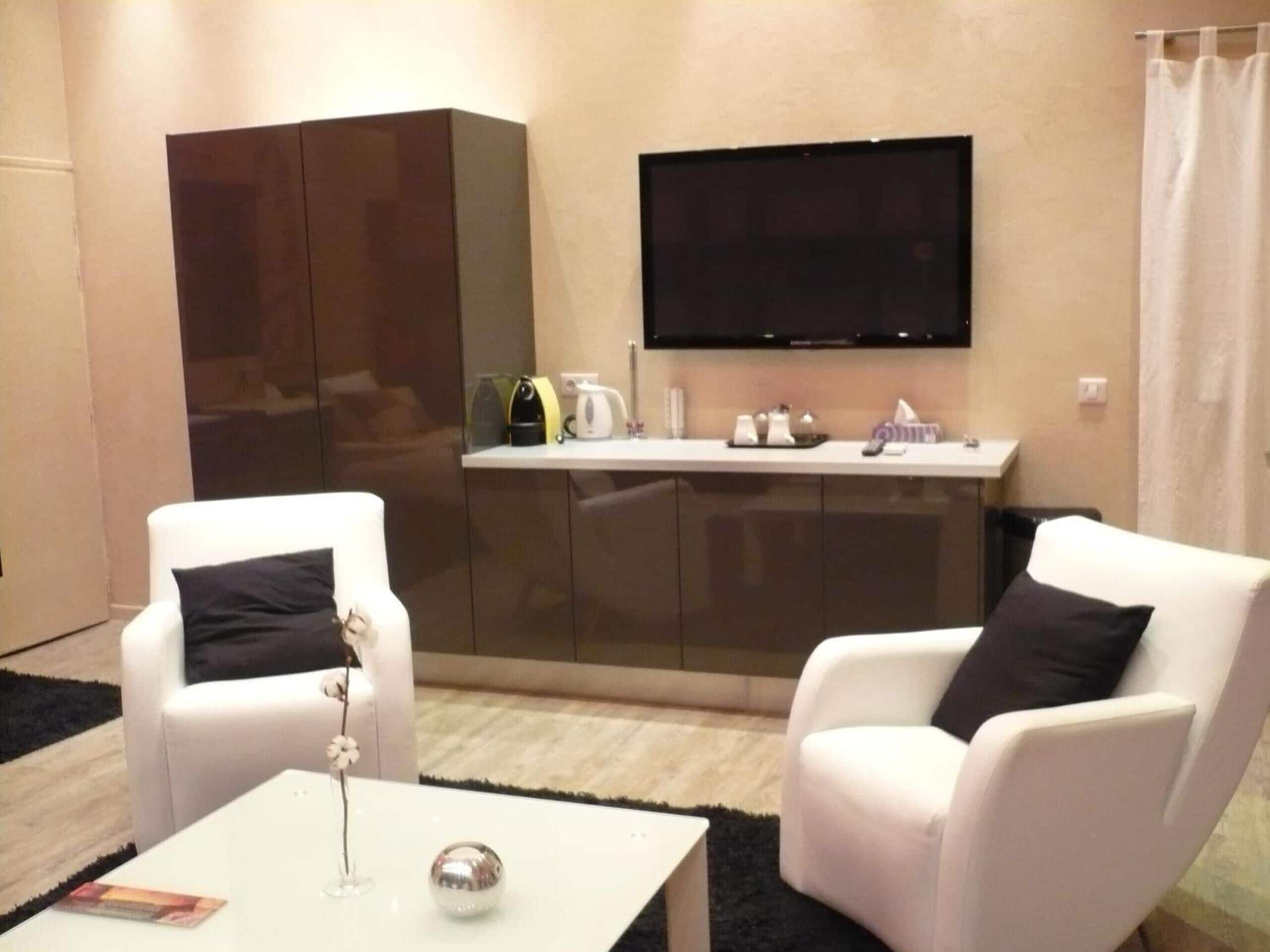 Een moderne woonkamer met twee witte fauteuils, een witte salontafel, een flatscreen-tv aan de muur en een glazen kast met keukengerei.