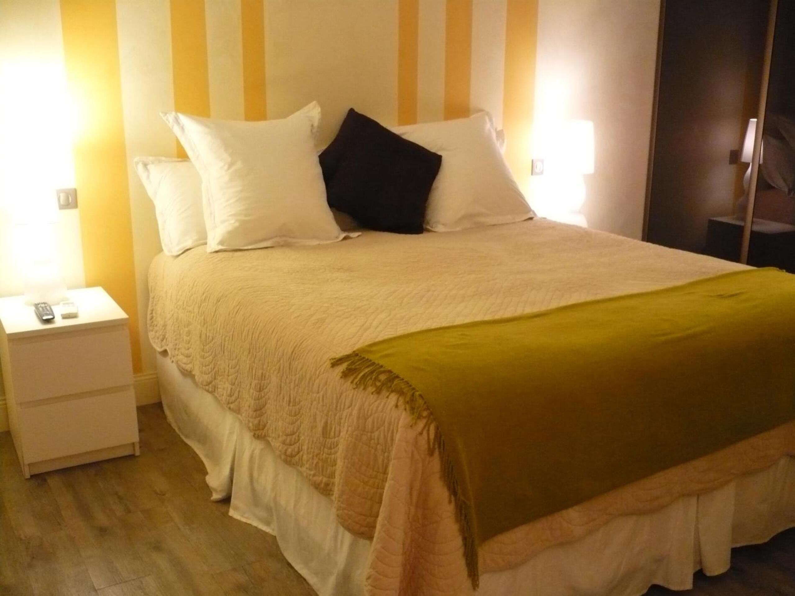 A neatly made bed with a white and black pillow arrangement, a green throw blanket, and a wooden nightstand with a remote control.