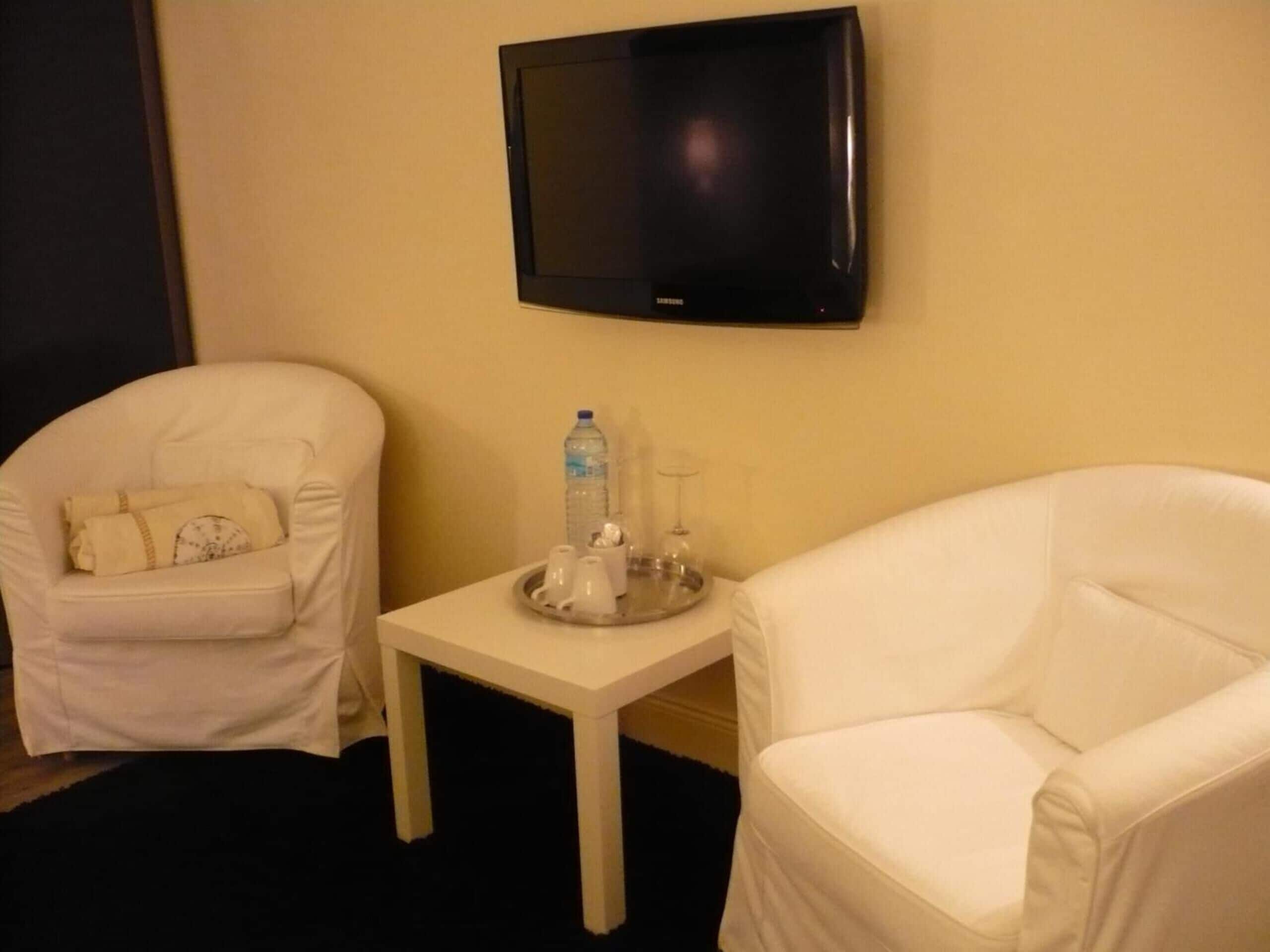 Une chambre d’hôtel équipée d’un téléviseur à écran plat fixé au mur, d’une petite table blanche sur laquelle se trouve un plateau avec des bouteilles d’eau et des verres, et de deux fauteuils blancs.