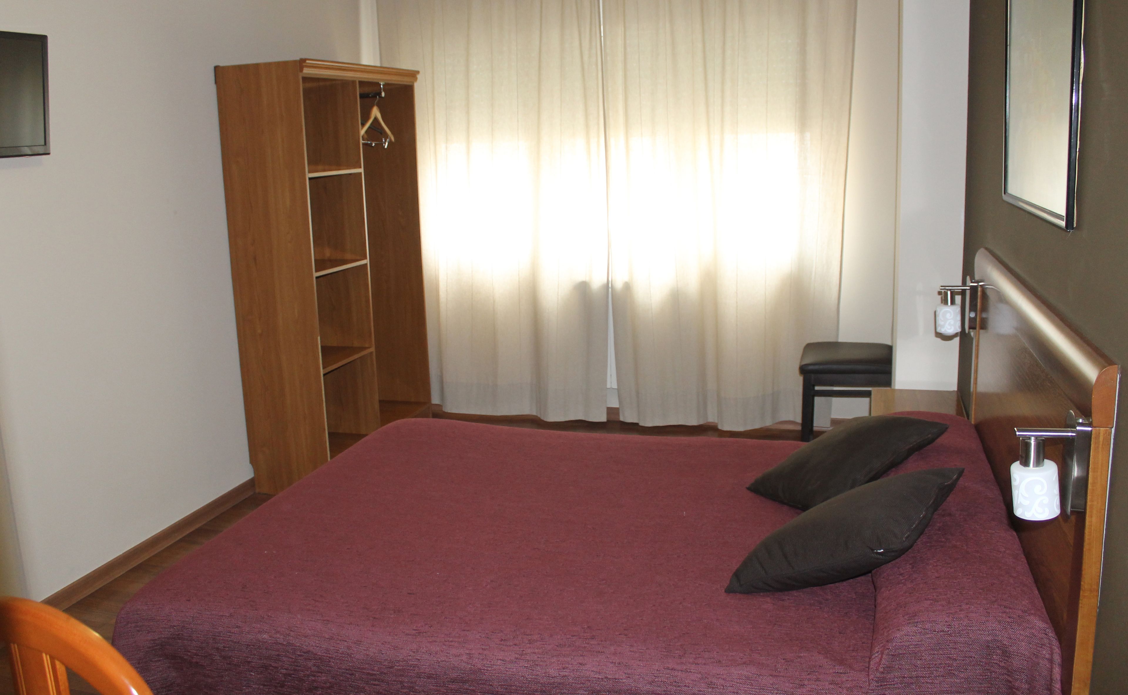 A hotel room with a bed, a wooden wardrobe, a small table, and a chair.