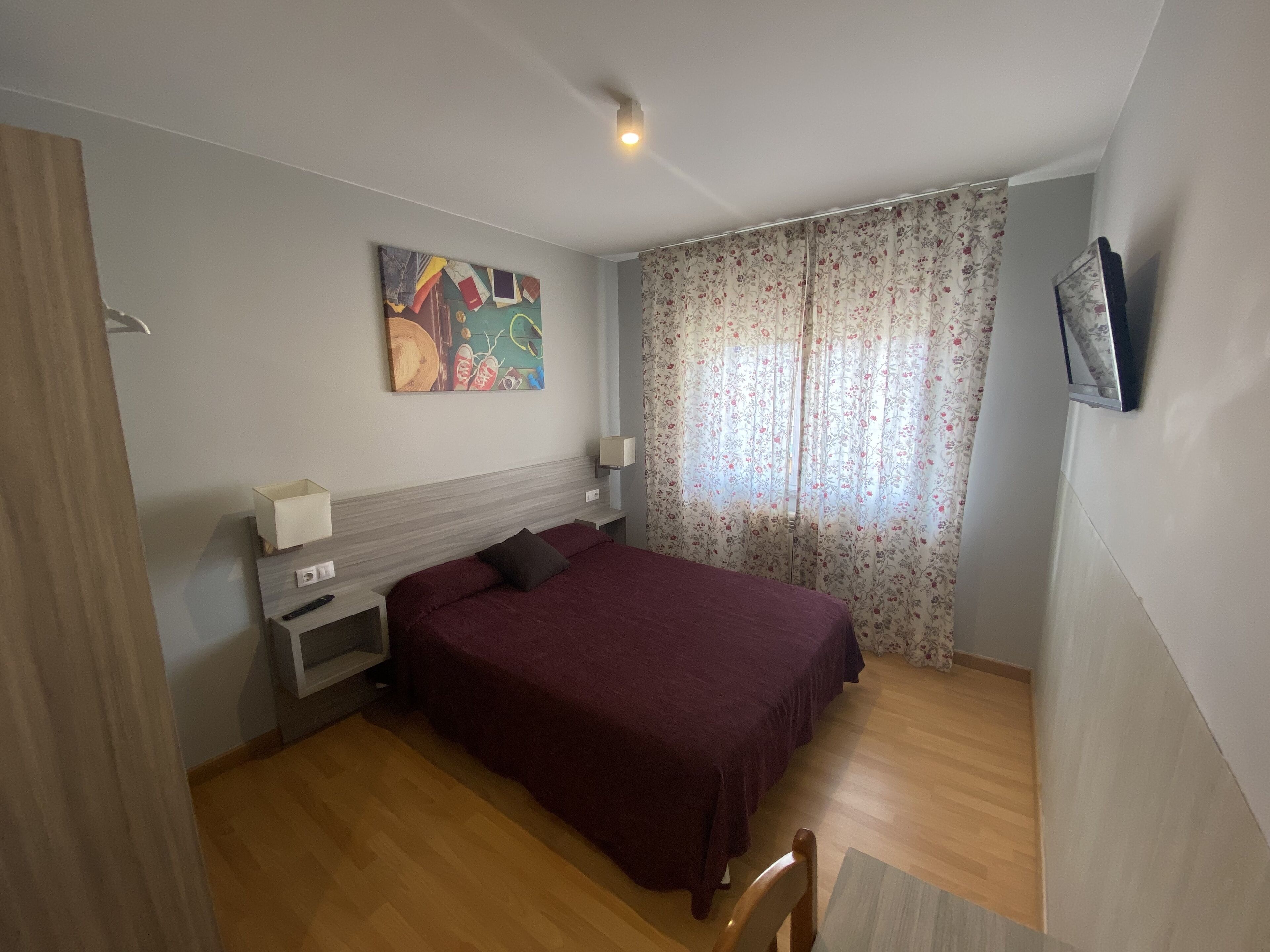 Un dormitorio con cama, mesita de noche, televisor montado en la pared, un cuadro y una ventana con cortinas florales.
