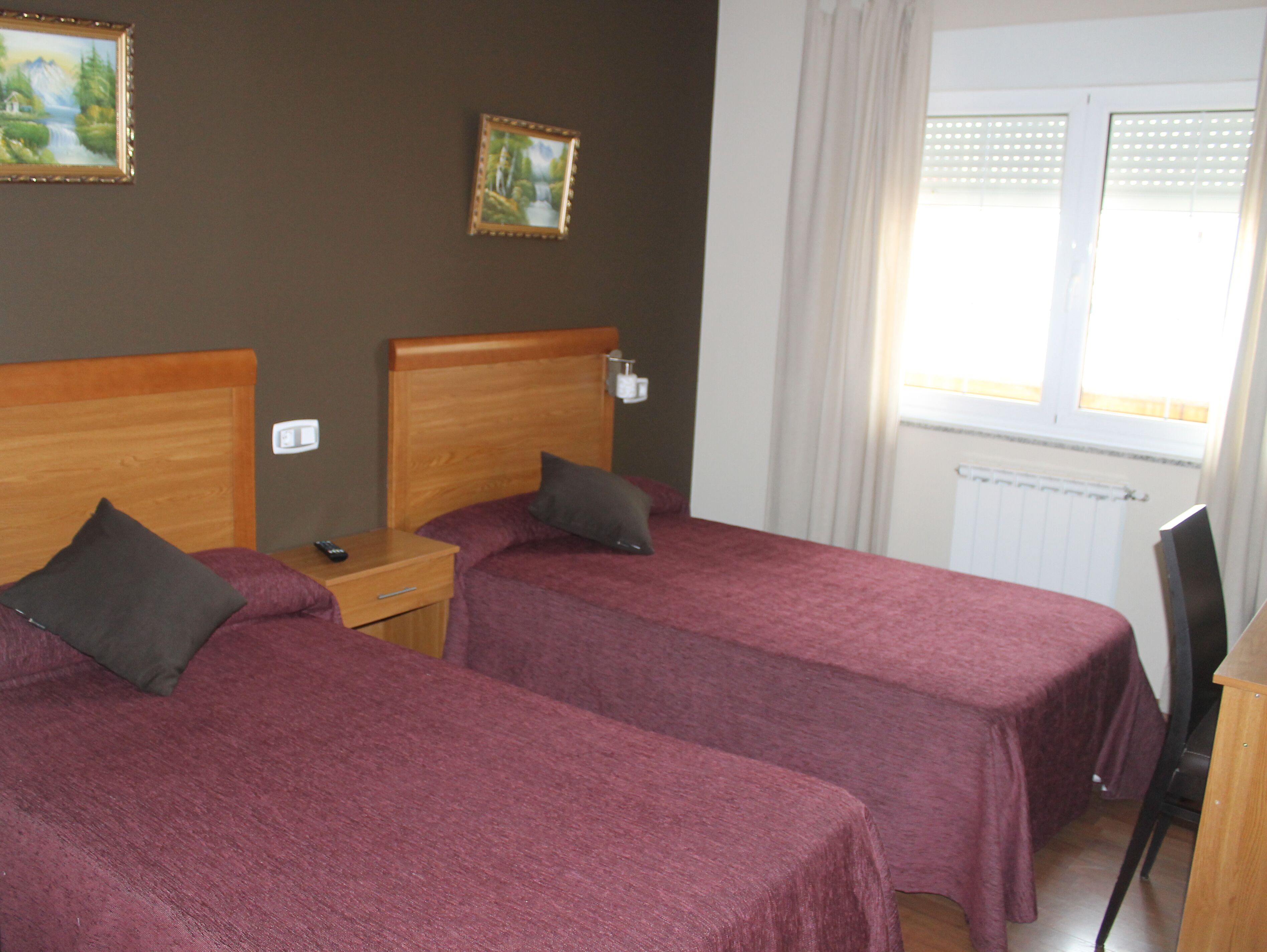 A hotel room with two single beds, a nightstand, a chair, a window with curtains, and a radiator.