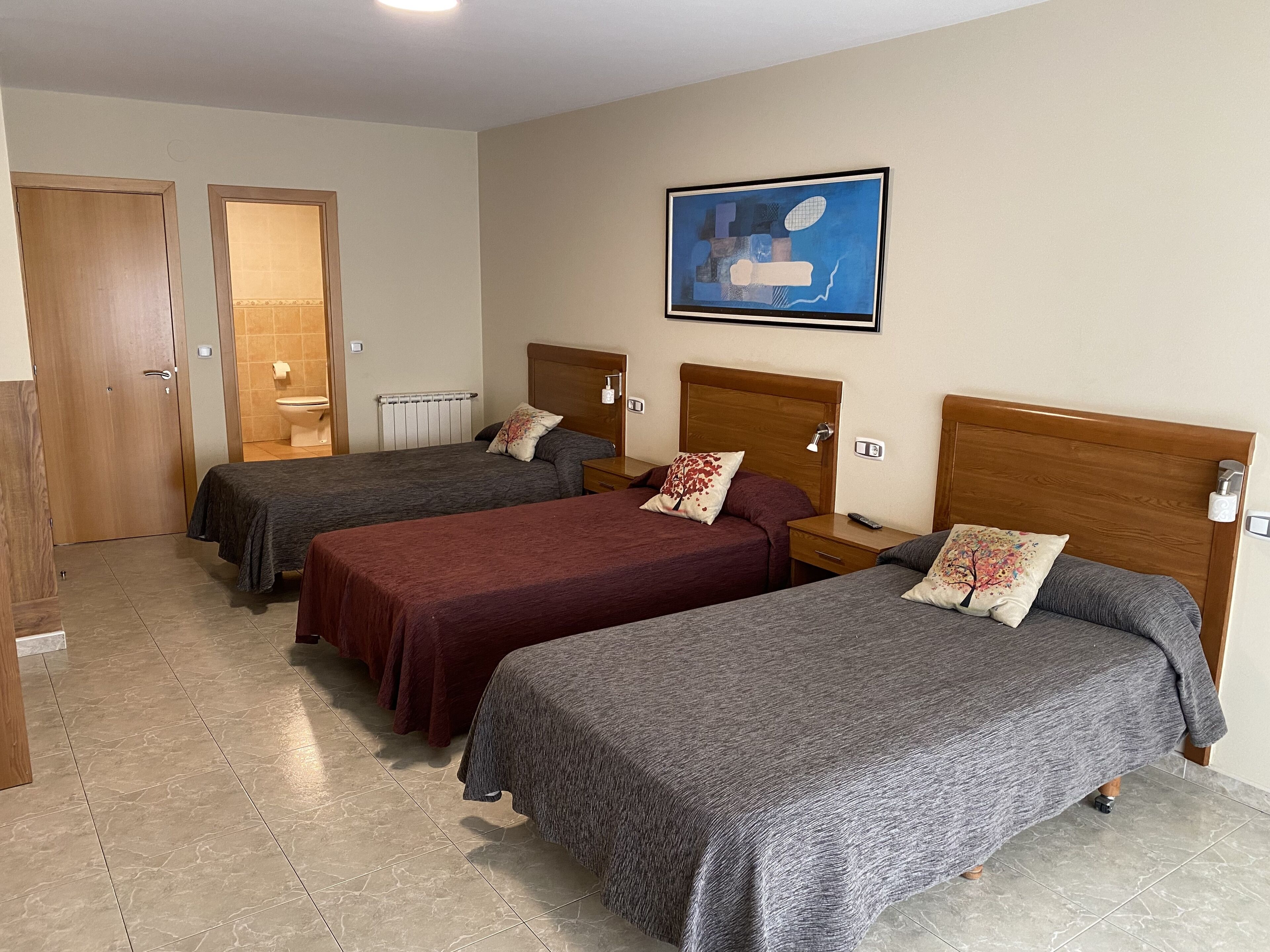 A hotel room with three single beds, a wooden door, a radiator, and a framed abstract painting on the wall.