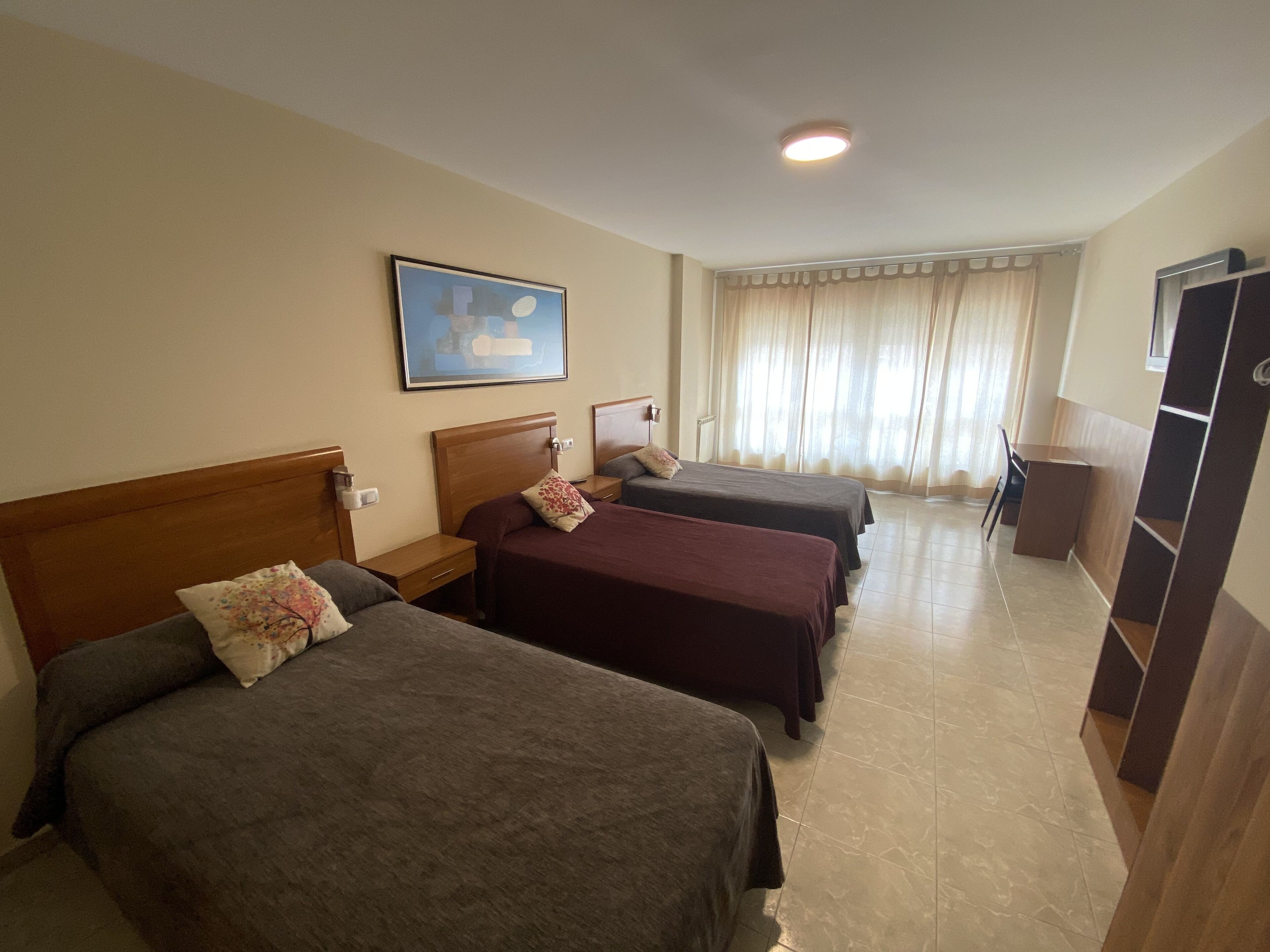 A hotel room with three beds, a painting on the wall, a window with curtains, and a desk with a chair.