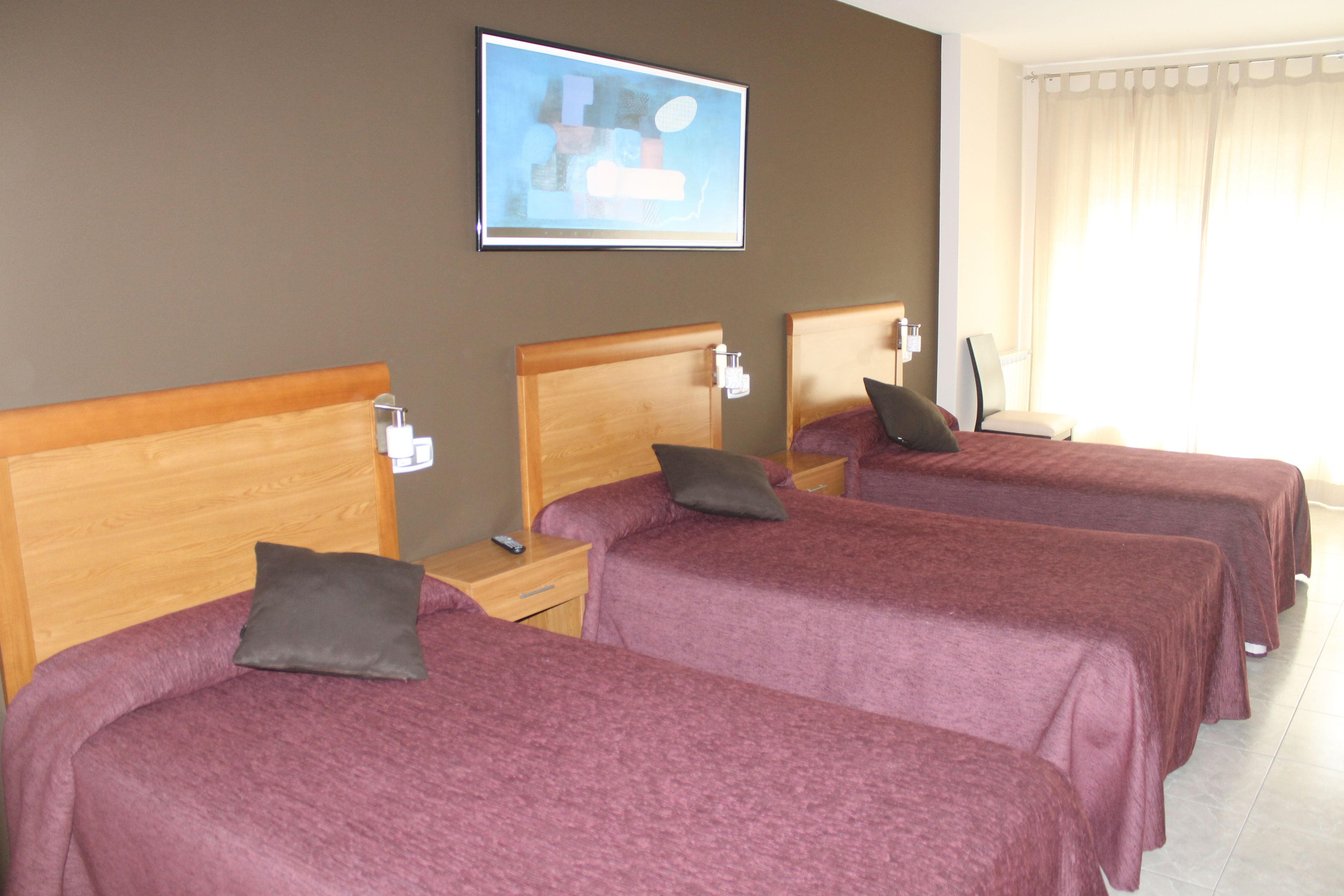 Habitación de hotel con tres camas individuales, cada una con cabecero de madera y colcha morada. Hay un cuadro enmarcado en la pared sobre las camas y una ventana con cortinas a la derecha.