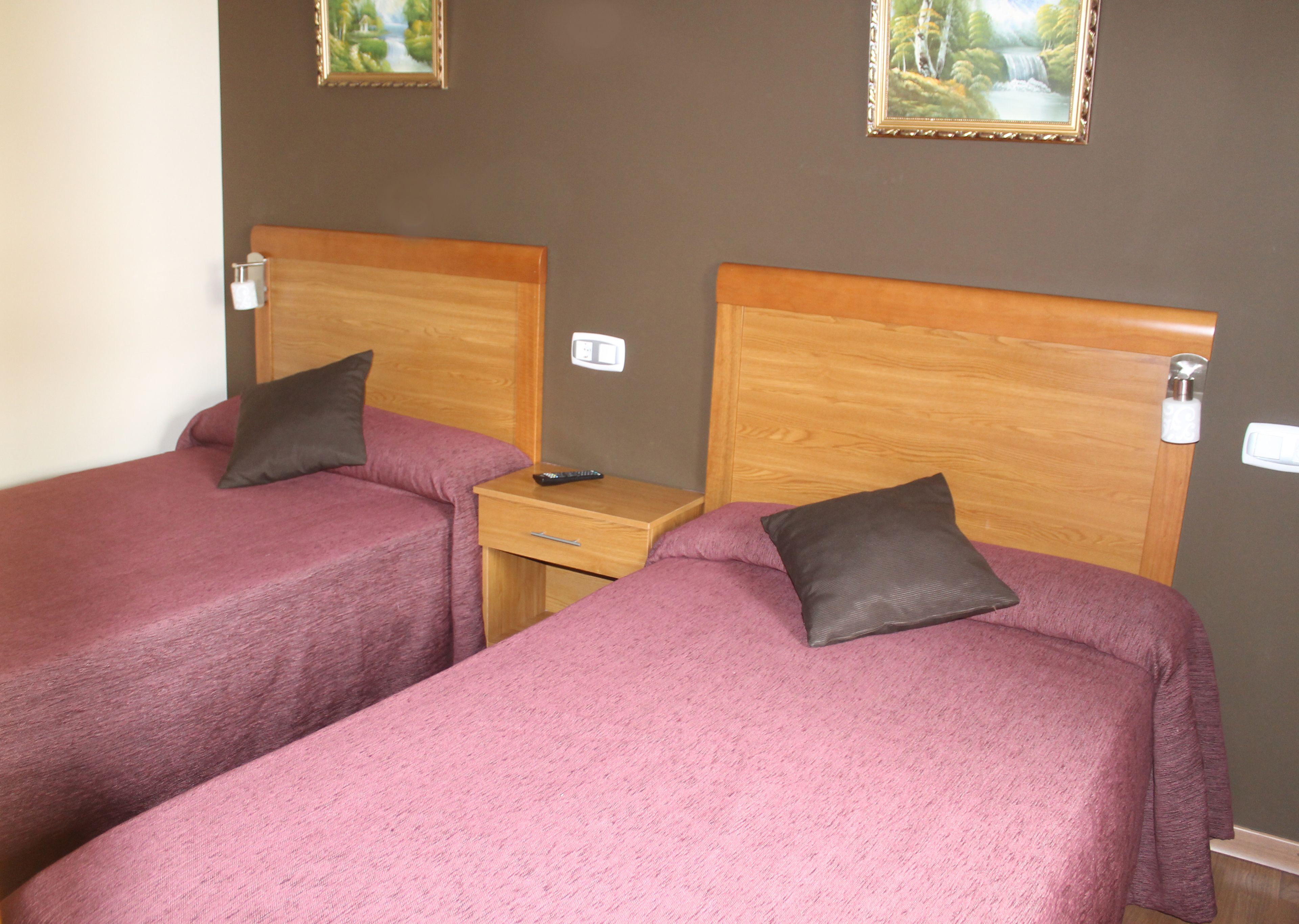 A hotel room with two single beds, each with a wooden headboard and a purple bedspread. There is a wooden nightstand between the beds with a remote control on it.