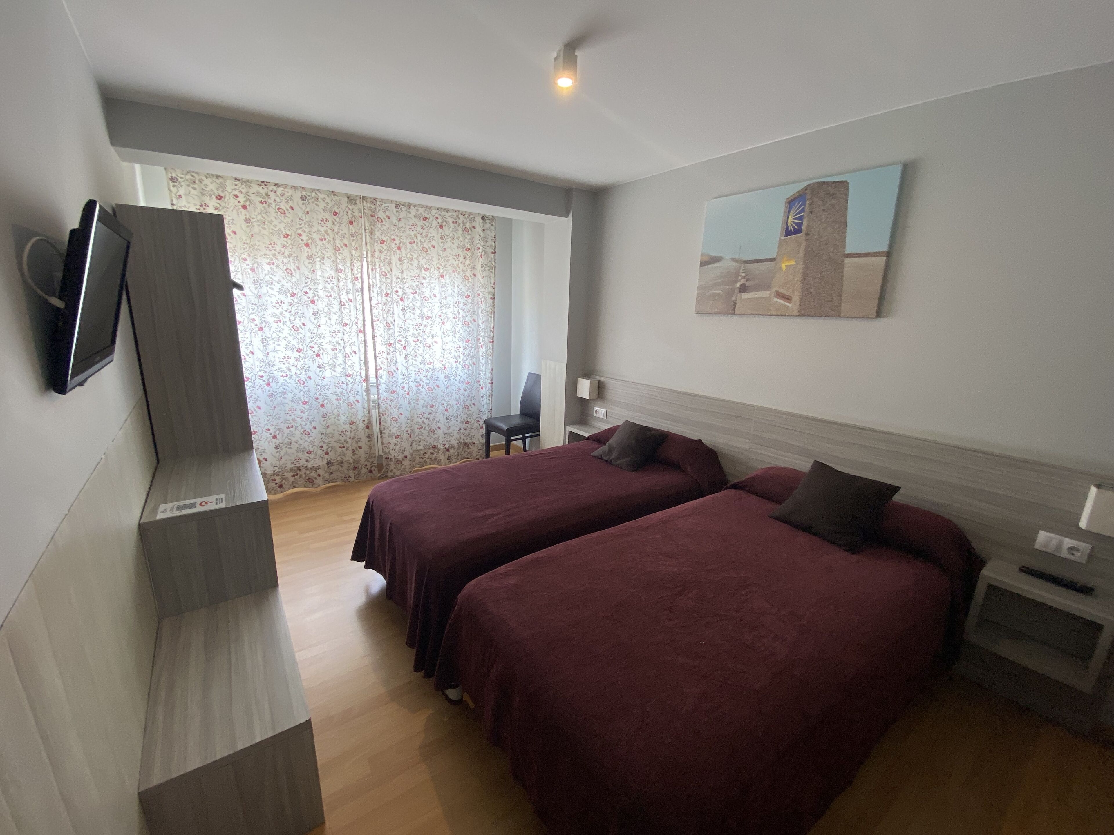 A hotel room with two beds, a flat-screen TV mounted on the wall, a painting on the wall, and a window with floral curtains.