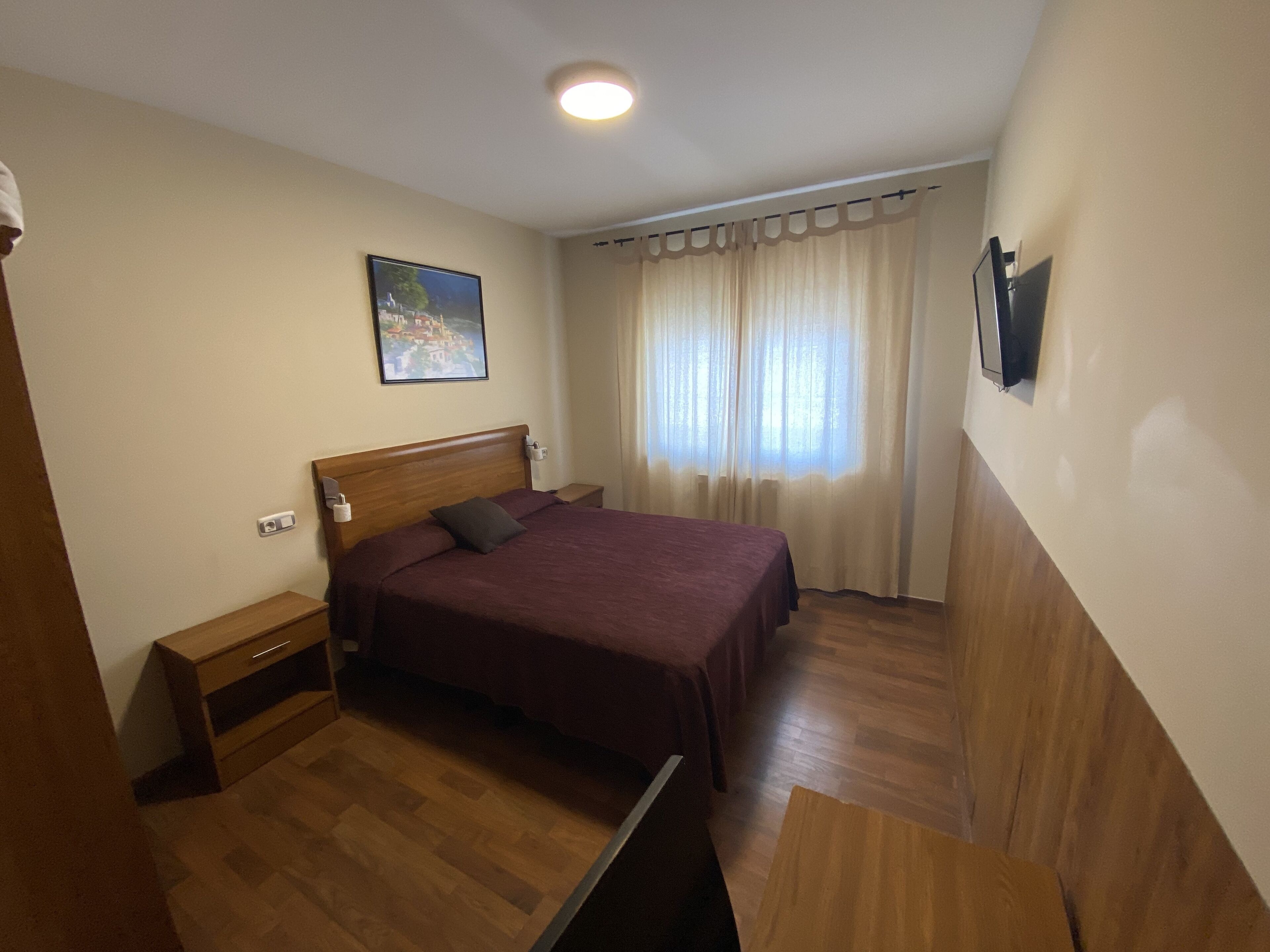 A bedroom with a wooden bed, a nightstand, a bedside table, a wall-mounted TV, and a window with curtains.