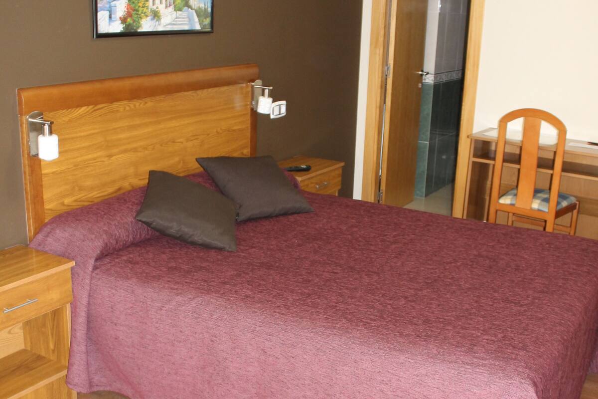 A bedroom with a wooden headboard, a bed with a purple bedspread, two bedside tables with lamps, a wooden chair, and a painting on the wall.