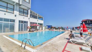 Zona de piscina con sillas de descanso y sombrillas, un flotador salvavidas y un edificio con un letrero que dice "CAFE BAR".