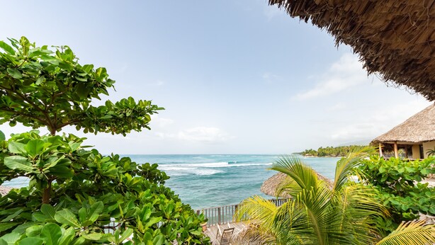 A wooden deck with chairs overlooking the ocean, surrounded by lush greenery and a thatched-roof structure. - Yoset Hotel (Cayes-Jacmel)