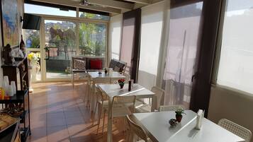 A dining area with white tables and chairs, a sofa with red cushions, and a view of a cityscape through large windows.