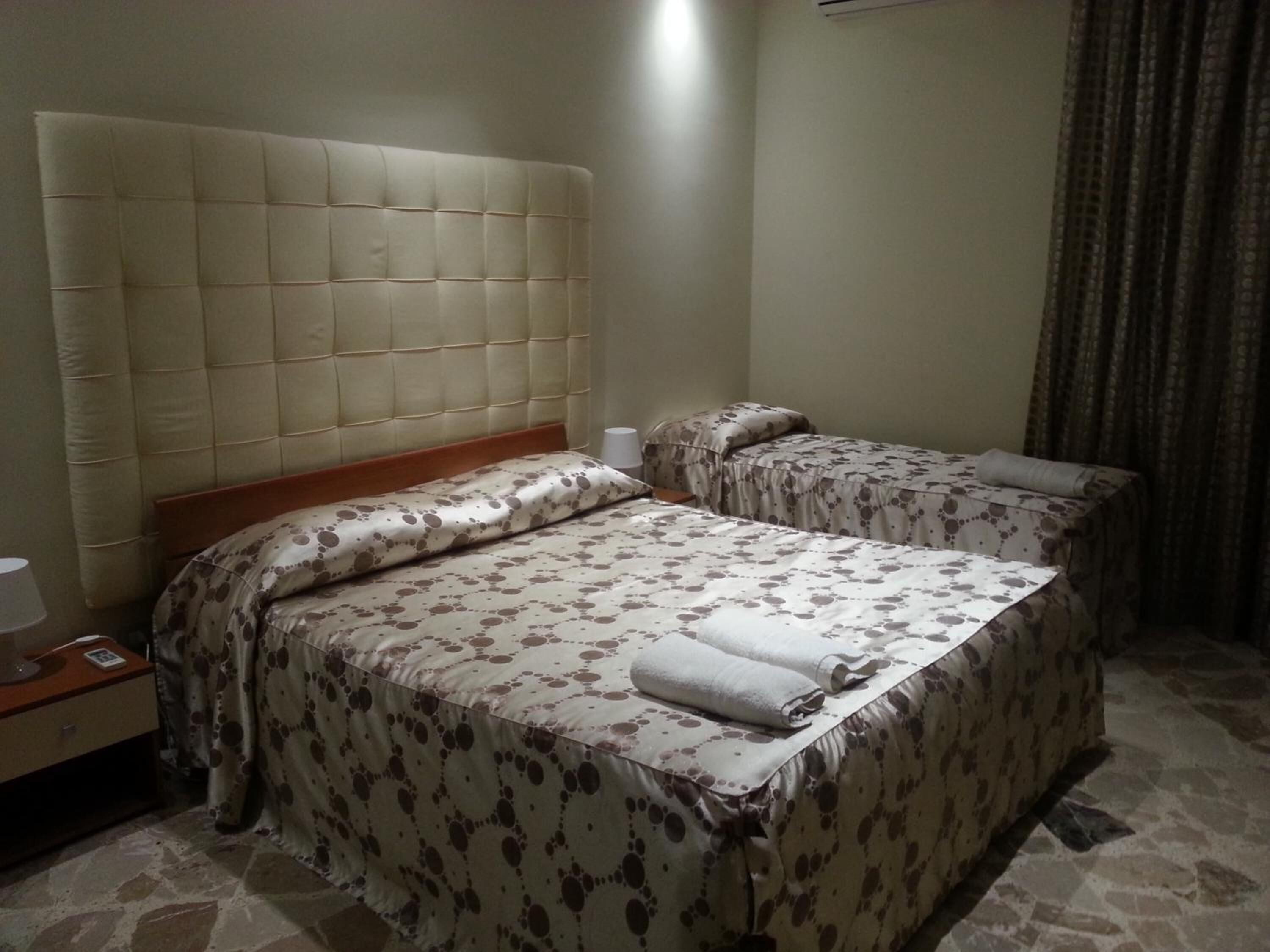 A hotel room with a large padded headboard, two beds with floral-patterned bedding, a nightstand with a lamp, and a curtain on the window.